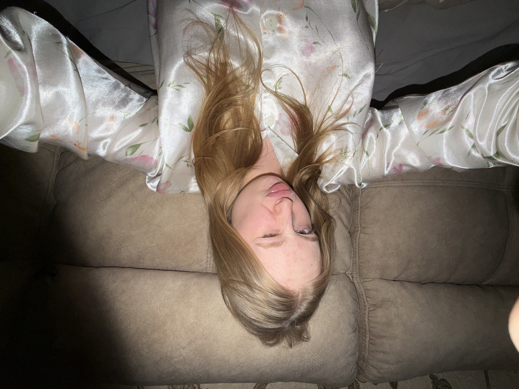 A female with fair skin, blonde hair, and wearing a floral-patterned satin pajama top lies on a beige fabric couch. She is positioned with her head at the bottom of the frame and winking her left eye. The background consists of the couch and parts of the pajama top. No information in the image indicates this is in Penza, Russia.
