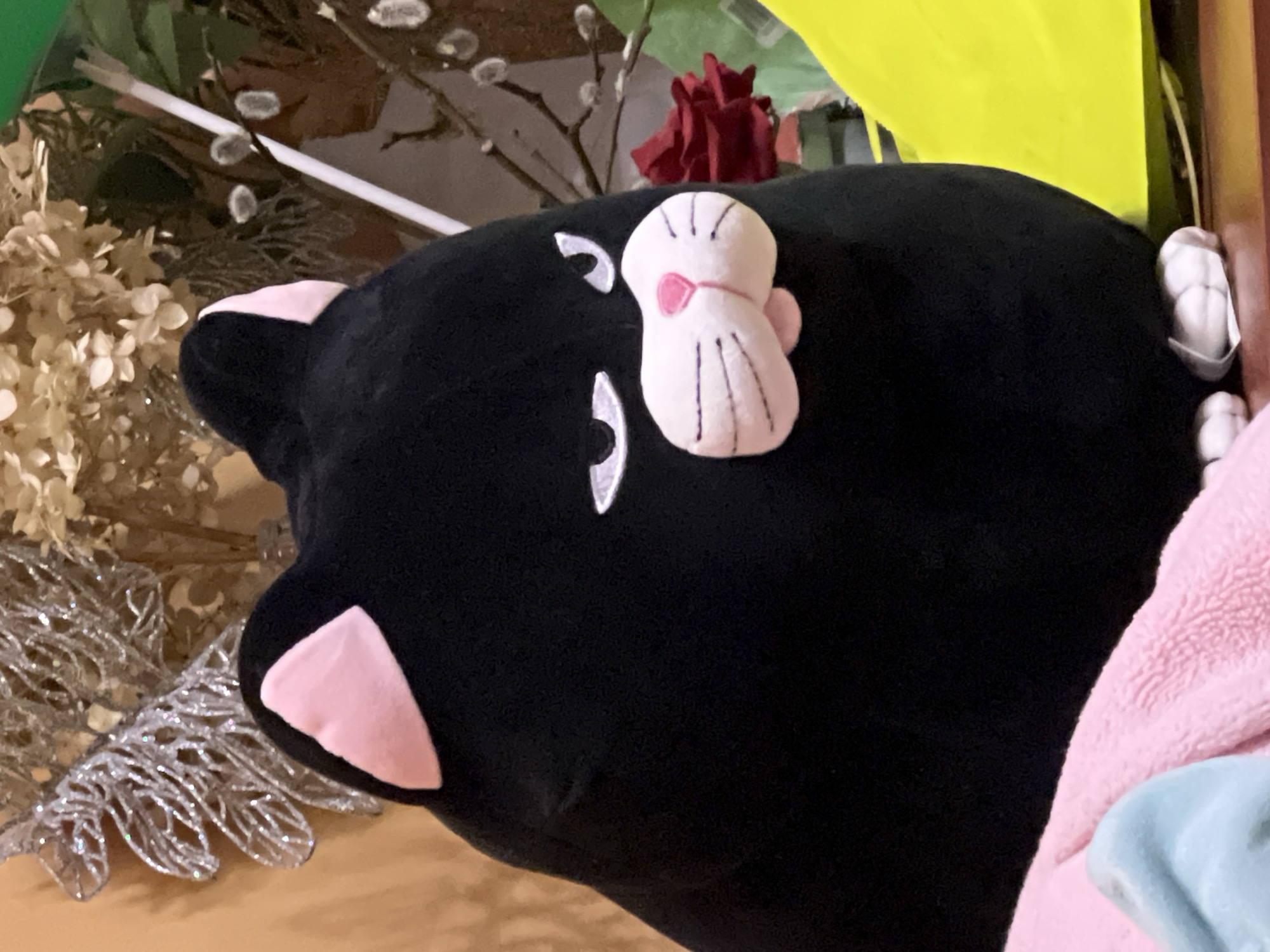 The image displays a plush toy cat with black fur and pink ears, sitting on a pink blanket and a blue cloth. The cat has a distinctive facial expression with white, angry-looking eyes and a pink nose with whiskers. In the background, there are decorative elements including a silver glittery branch-like ornament and dried flowers. A red rose is visible to the right of the cat. The scene appears to be indoors, possibly in a residential setting, but no specific location in Minsk, Belarus can be determined from the visual information provided. The lighting is warm and suggests an indoor environment.