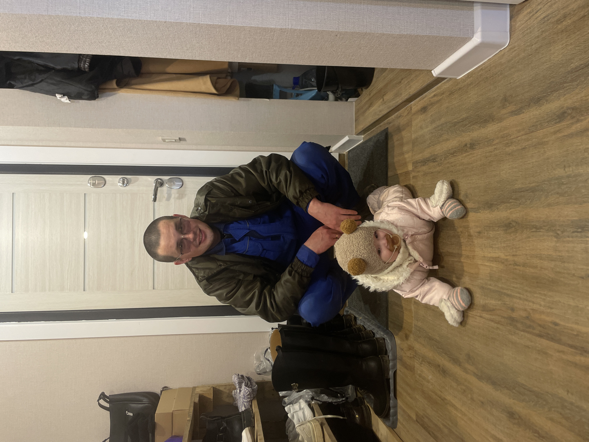 An indoor photograph, taken in Подшивалово, Russia, captures a man and a baby in what appears to be a hallway or entryway. The image is oriented vertically (portrait mode), with the man crouching on the left side and the baby lying on its stomach on the right.
The man, an adult with short dark hair and glasses, is wearing a dark olive-green jacket over blue work overalls. He is smiling broadly and looking directly at the camera. His right arm is bent, and his left hand is placed near his knee.
Beside him, a baby or young child is dressed in a light pink quilted snowsuit with a white fur trim around the hood. The child also wears a light brown knit hat with two small, fuzzy bear-like ears. The baby is lying face down, propped up slightly, and is looking up towards the camera with a curious expression, possibly with a pacifier partially visible in its mouth. Fuzzy grey and white striped slippers are visible on the baby's feet.
The floor is made of light brown wooden planks. In the lower right corner, several pairs of dark-colored boots are neatly arranged on a grey mat, alongside other shoes on a wooden rack. A black backpack or bag is visible on the floor to the left of the footwear. The background features a light-colored wooden door with multiple silver handles and locks. Above the door and along the upper wall, various items of clothing and other objects are stored in an open cabinet or shelf. A white cable management conduit runs along the wall.
The scene suggests a warm, domestic moment, possibly as the baby is preparing to go out or has just returned from cold weather, given its attire. There is no visible text in the image.