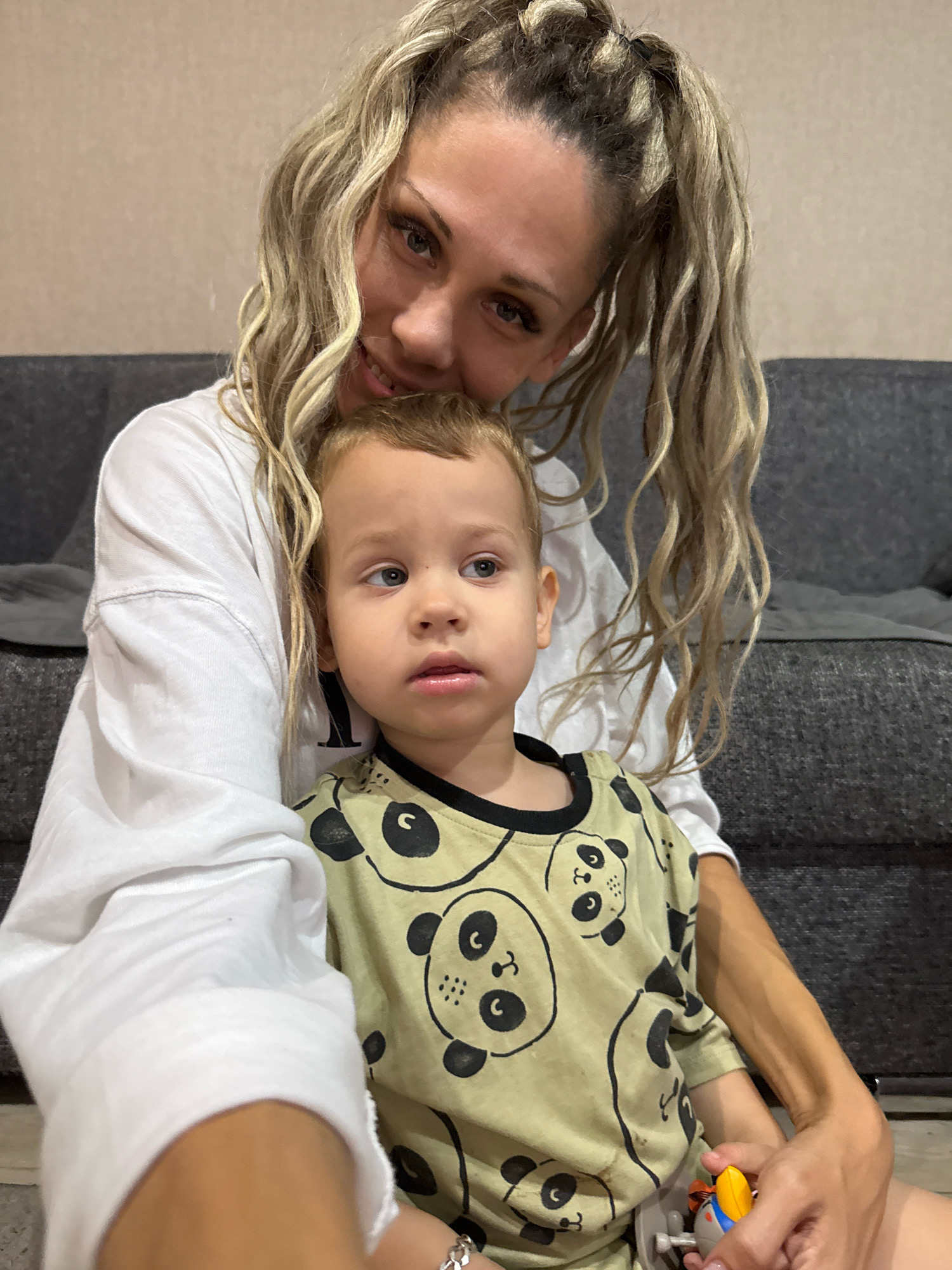 A young woman with blonde, pigtail-styled hair and heavy eye makeup is holding a toddler boy in her lap. The woman is wearing a white long-sleeved shirt. The toddler is wearing a light green shirt with a panda print. The two are seated on the floor in front of a dark gray couch. In the toddler's hands is a small toy, partially visible. The background wall is a neutral beige color. The image is a selfie, taken from the woman's perspective.