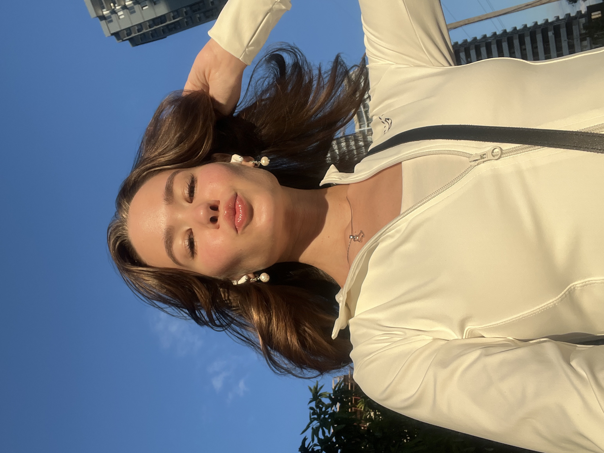 A young woman with brown hair is taking a selfie outdoors against a bright blue sky. She is wearing a white zippered jacket over a white tank top and a necklace. She has on pearl earrings and is puckering her lips, with her right hand touching her hair. In the background, to the left, is a leafy tree, and to the right are tall buildings and utility poles, suggesting an urban environment. The lighting indicates it is daytime, possibly late afternoon due to the warm glow on her face. The location is the city of Batumi, Georgia.