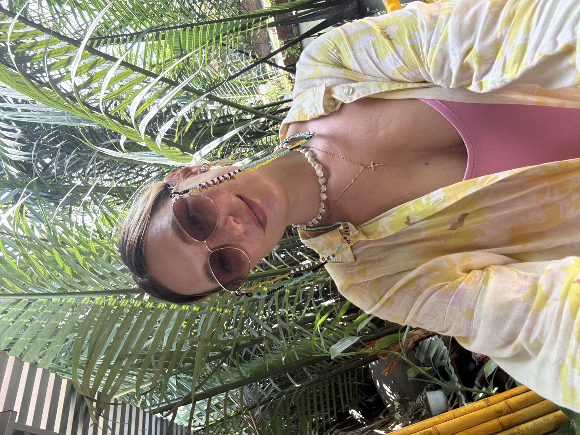 A woman is captured in a selfie, smiling and looking towards the camera. She is outdoors, with lush green palm fronds forming the backdrop, suggesting a tropical setting. The light suggests it is daytime. She wears sunglasses with beaded chains, a pearl necklace, and a delicate gold cross pendant over a pink top and a yellow and white floral print shirt. Her left arm rests on a bright yellow bamboo structure. The scene appears to be in a garden or resort area, possibly in Đà Nẵng, Vietnam, given the prominent bamboo element and tropical foliage.