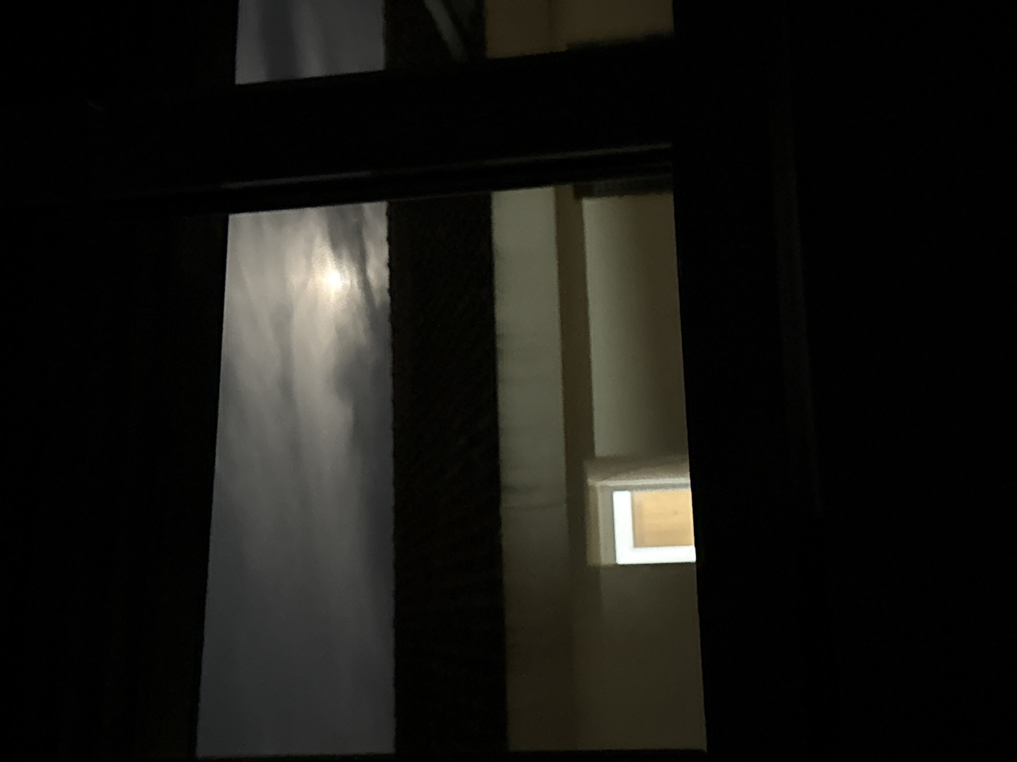 A view through a window at night. The window frame is dark and obscures most of the scene. On the left, a blurred white or gray shape suggests a bright object, potentially the moon or strong light source, seen through a translucent material. Through a portion of the window, an interior room is visible, illuminated with a warm light, possibly from a lamp or internal light source. A small window inside the interior is partly visible with a lit opaque window. The lack of detail and clarity makes it difficult to definitively identify the location or any other objects or people.
