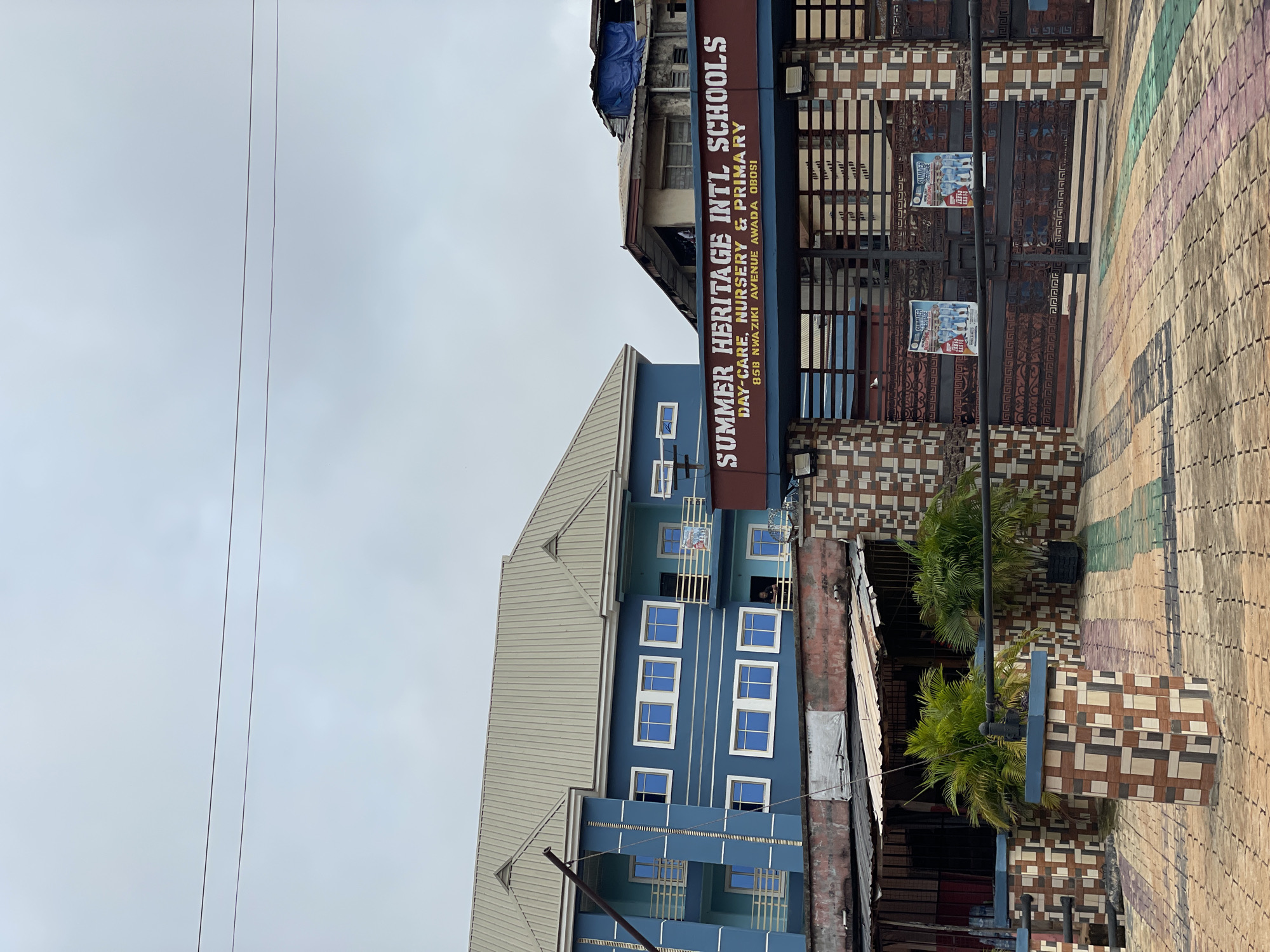 The image shows a street scene in Onitsha, Nigeria, featuring the "Summer Heritage Intl Schools," a blue building with a beige roof and white-framed windows. A sign adjacent to the school building states its name, "Summer Heritage Intl Schools," along with "Day-Care, Nursery & Primary," and its address: "85B NWAZIKI AVENUE AWADA OBOSI." Power lines stretch across the overcast sky. A decorative brick pillar and patterned walkway border the building, while a gated entrance with posters is visible on the right. Two small palm trees are situated near the entrance.