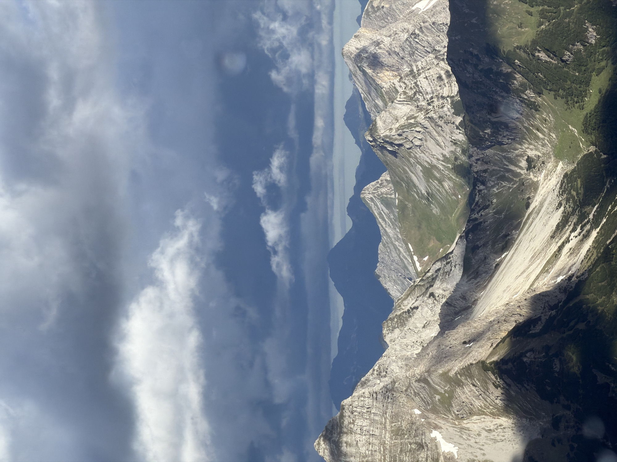 The image depicts a panoramic view of the Alps, likely near Grainau, Germany, under a dramatic, cloudy sky. The foreground is dominated by rugged, rocky mountain peaks with sharp ridges and steep slopes. Patches of snow are visible on some of the higher elevations, contrasting with the grey rock and green vegetation lower down. The midground features more distant mountain ranges, appearing in softer focus and shrouded in a bluish haze. The sky is filled with dark, heavy clouds, with some lighter, scattered clouds interspersed, suggesting a dynamic weather system. Sunlight breaks through in places, casting dramatic shadows across the mountain faces and highlighting the textures of the landscape. The scene conveys a sense of vastness and the raw beauty of alpine terrain.