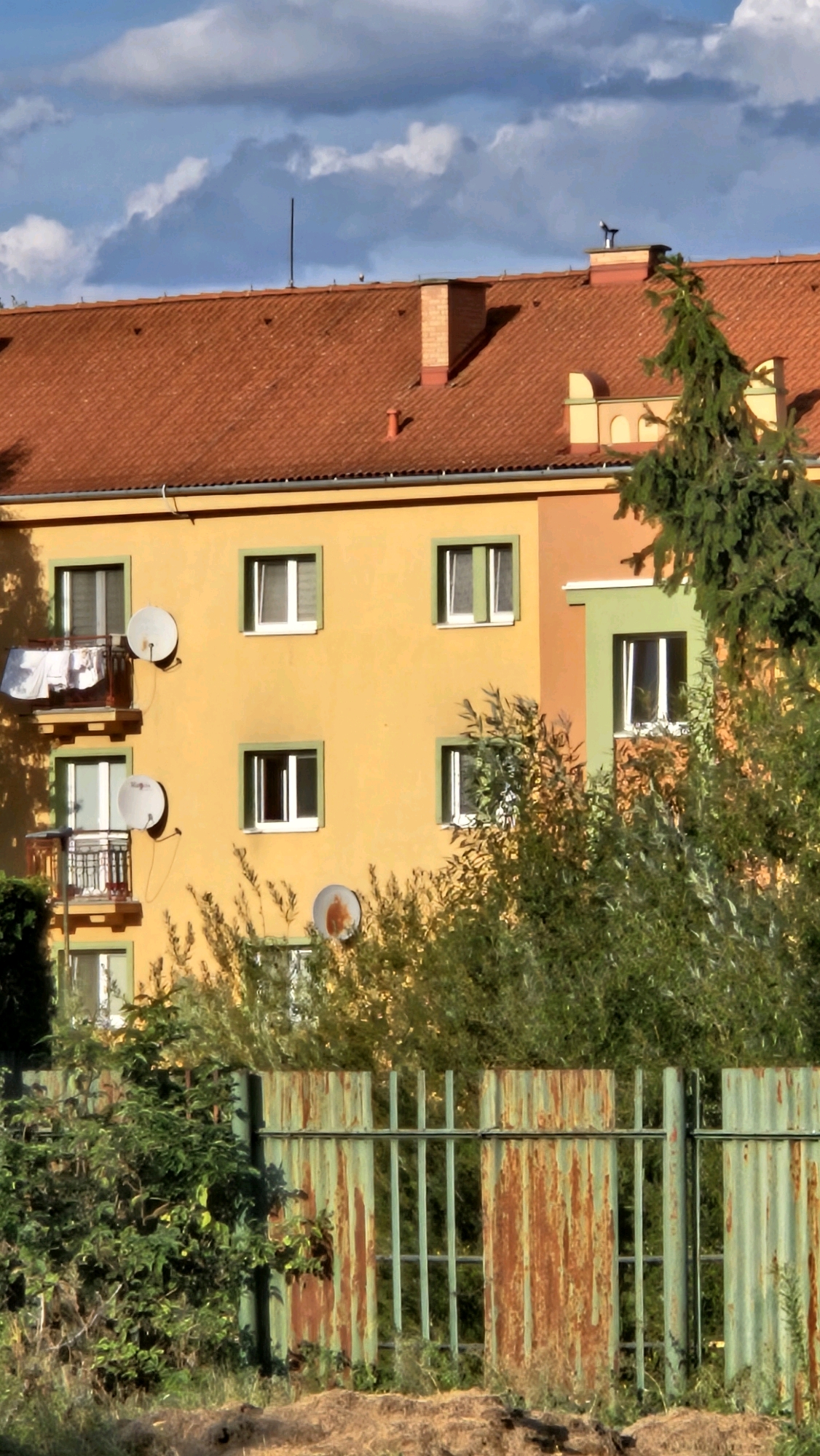 The image depicts a residential building with an orange facade and a red tiled roof under a partly cloudy sky. The building features several windows, some with green frames, and balconies with satellite dishes. Laundry is visible hanging on a clothesline on one of the balconies. In the foreground, a rusty green corrugated metal fence is present, with overgrown vegetation behind it and in front of the building. The scene is likely a view of an apartment building in a residential area of Prešov, Slovakia.