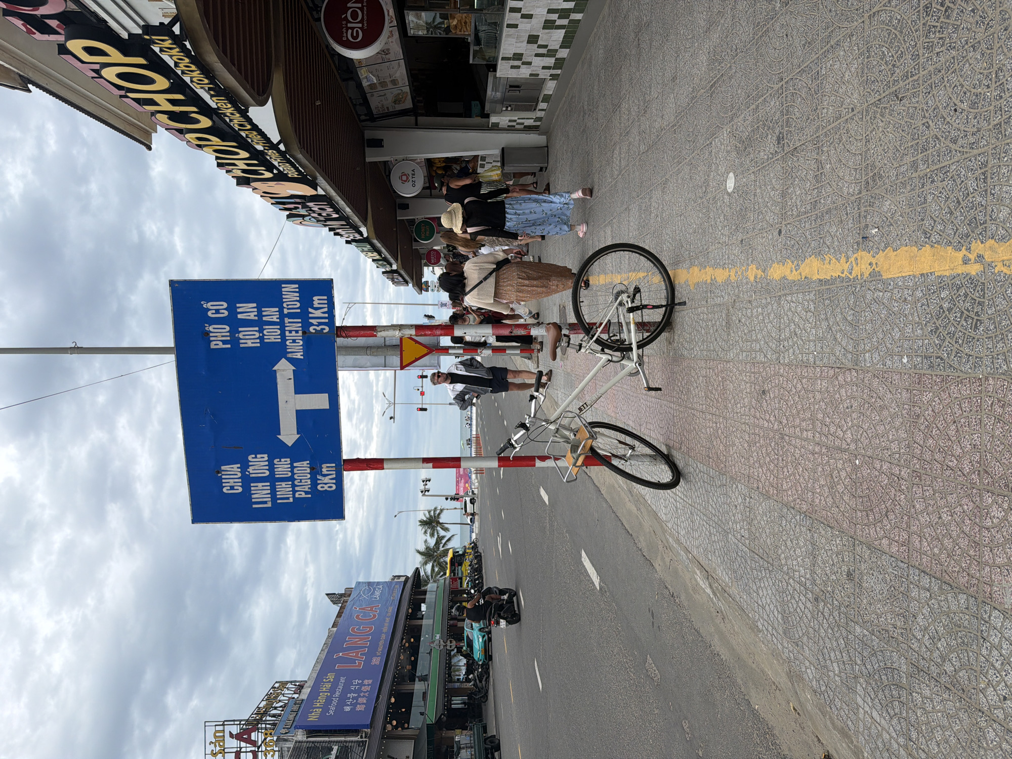 In Đà Nẵng, Vietnam, a white bicycle is parked on a sidewalk paved with patterned tiles. A blue street signpost is located next to the bicycle. The sign indicates directions to "CHUA LINH UNG" and "LINH UNG PAGODA" with an arrow pointing left, stating "8Km." It also indicates directions to "PHO CO HOI AN" and "ANCIENT TOWN" with an arrow pointing right, stating "31Km." Several people are walking on the sidewalk. On the street, a motorbike is being ridden by a person wearing a helmet. In the background, there are buildings with signs, palm trees, and a cloudy sky.