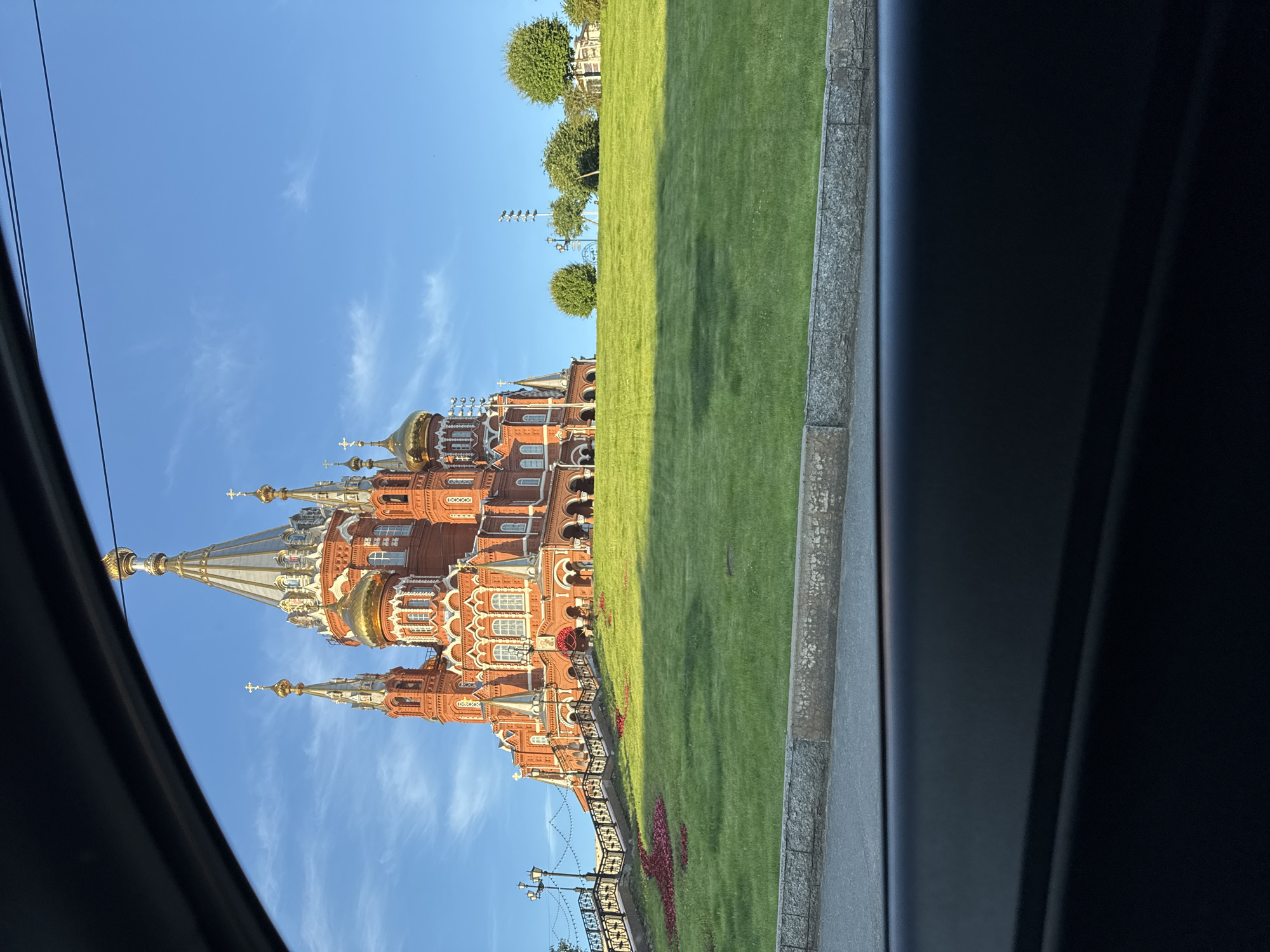 The image captures a wide shot of the Alexander Nevsky Cathedral in Izhevsk, Russia, on a clear, sunny day. The cathedral, constructed of red brick with white architectural accents, dominates the upper right portion of the frame, its multiple golden domes and spires gleaming under the blue sky. The building is situated atop a lush, green grassy hill. A decorative element, possibly made of red flowers, is arranged in a pattern on the grass in front of the cathedral's steps. A stone retaining wall and a paved area are visible at the bottom of the hill, with what appears to be a road or sidewalk beyond. The foreground is partially obscured by the dark interior of a vehicle, suggesting the photo was taken from inside a car. The overall scene is tranquil and picturesque, highlighting the grandeur of the cathedral and the surrounding park-like setting.