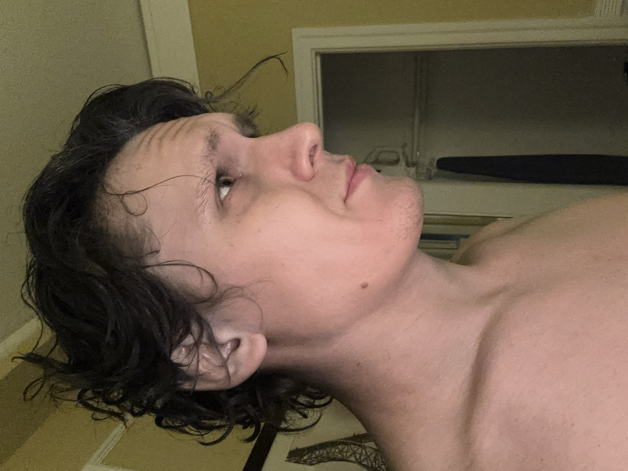 The image is a close-up, profile view of a person's head and upper torso, facing right. The subject has dark, wavy hair, light skin, and a mole on their left cheek. They appear to be indoors, in front of a yellow wall. A doorway with a dark object hanging inside is visible in the background, along with a framed object on the left wall. The lighting is dim. There is no indication of the location being San Francisco, United States of America.