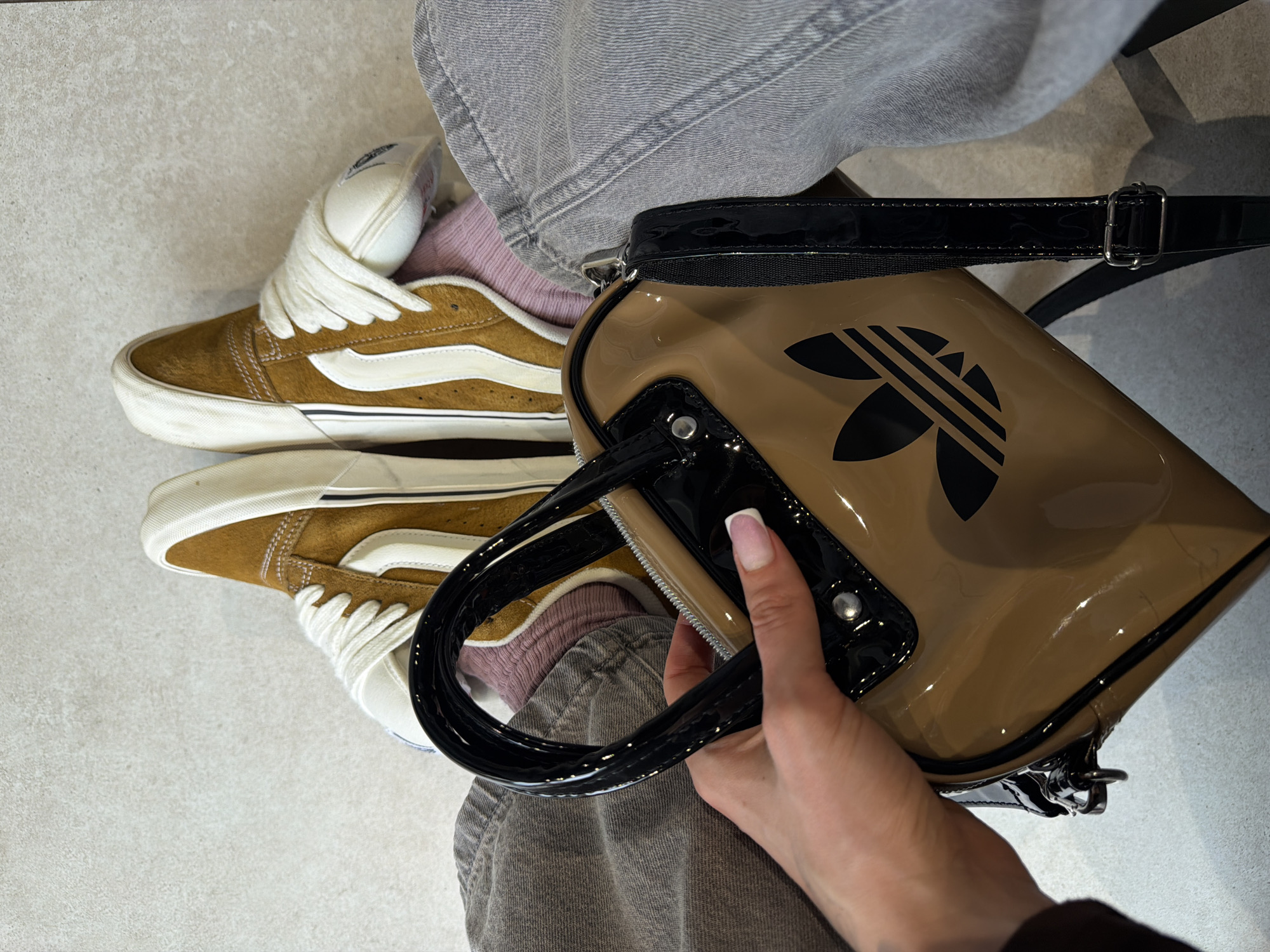 The image presents a close-up, high-angle view of a person's lower body, highlighting their footwear, clothing, and a handbag. The individual is wearing light gray, possibly denim, pants with visible seams, paired with light pink/purple crew socks. On their feet are brown suede Vans Old Skool sneakers, featuring white laces, a distinctive white sidestripe, and off-white soles that show slight signs of wear.
A hand, adorned with a French manicure, holds a glossy tan-colored handbag. The bag has prominent black patent leather accents, including its handles, trim, and an adjustable strap with a silver buckle. A large black Adidas Trefoil logo is clearly displayed on the front of the bag. The flooring beneath the person is a light, neutral color, possibly concrete or tiles, suggesting an indoor or well-lit outdoor setting. The location context for this scene is Budva, Montenegro. The time of day and specific weather conditions are not evident from the photograph.