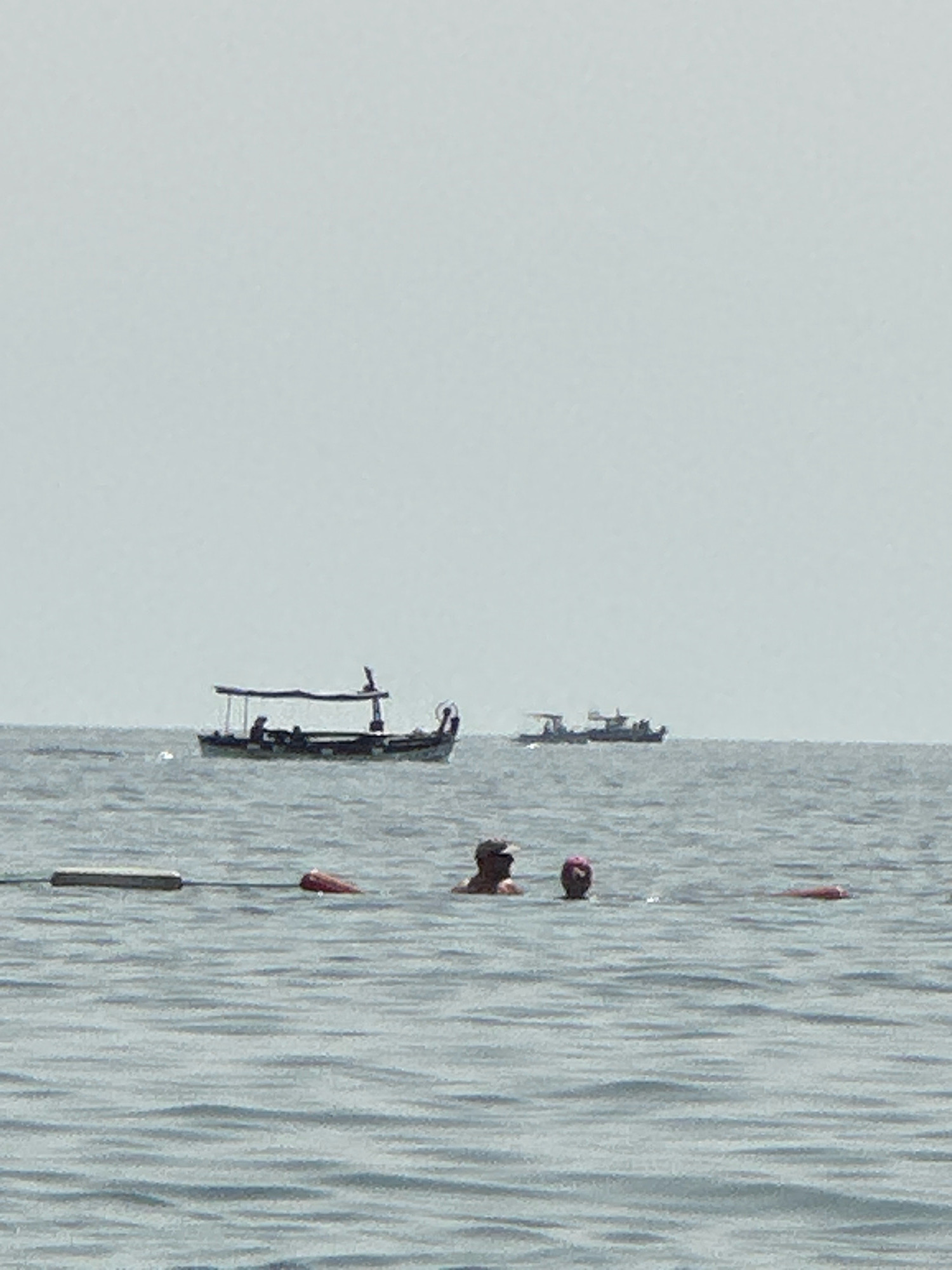 In the city of Boreti, Montenegro, the image captures a serene coastal scene. The vast expanse of the sea is the dominant element, with a slightly hazy sky overhead. In the mid-ground, two boats are visible. The boat on the left is a traditional wooden fishing vessel with a canopy. It appears to be stationary, with silhouettes of people on board. Further to the right, another boat is seen, also appearing to be at anchor. In the foreground, two individuals are partially submerged in the water, suggesting they are swimming or wading. One person, seen from behind, is wearing a dark cap. The other person, closer to the right edge, is wearing a pink cap. Several buoys or markers are also visible in the water, indicating a designated swimming area or a marine boundary. The overall atmosphere is calm and leisurely, indicative of a typical day at the beach.