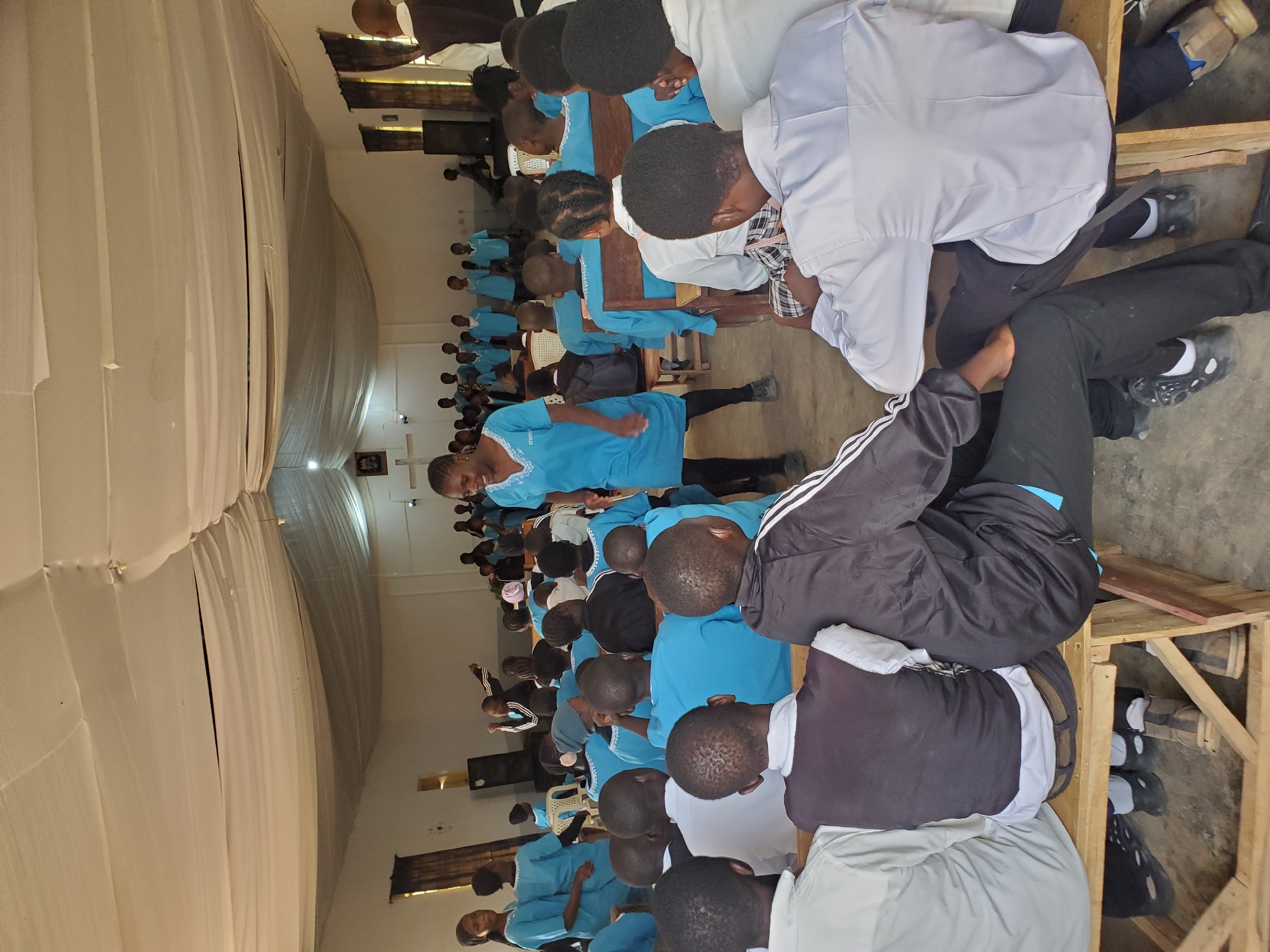The image depicts a gathering of people, likely in a hall or large room, in Kuru Karama, Nigeria. The subjects are primarily children and young adults, many wearing matching blue outfits, suggesting a school or religious event. They are seated on wooden benches and some chairs, facing towards the front of the room where a few adults are also present. Some individuals appear to be singing or engaged in a presentation, with hands raised. The atmosphere seems formal and organized.
The setting is characterized by simple wooden furniture and a draped ceiling, possibly made of fabric, creating a somewhat makeshift but functional space. The lighting appears to be artificial, indicating an indoor event. The clothing of the attendees, especially the blue uniforms, is a notable detail. Some individuals are also wearing casual wear like tracksuits. The overall mood seems to be one of communal participation and focus on the ongoing activity.
No specific time of day or weather conditions can be determined from the image. There is no visible text within the frame. The event appears to be a collective activity, possibly a service, performance, or meeting, with a strong sense of unity among the participants.
