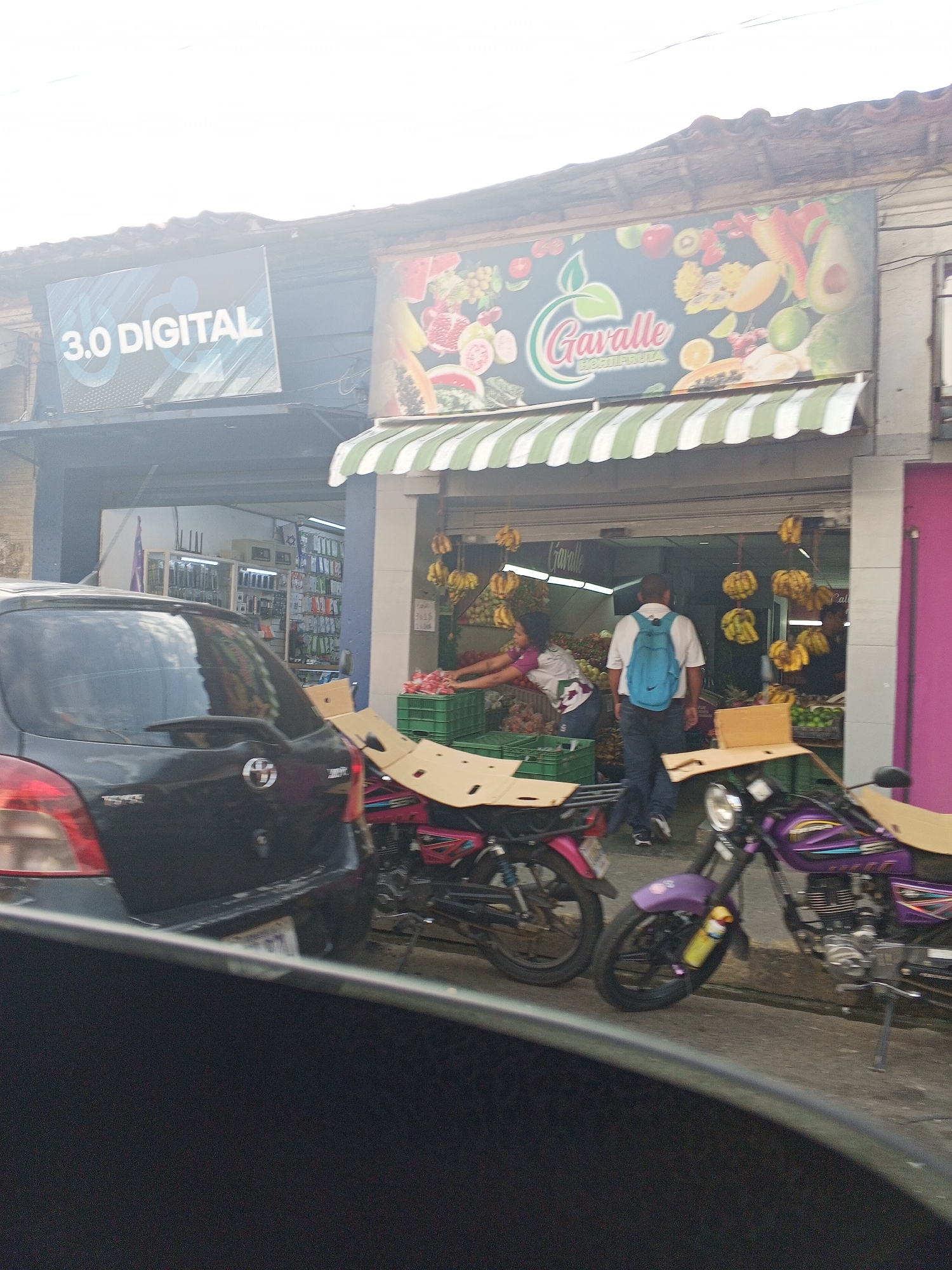 The image depicts a street scene in Tinaquillo, Venezuela. A black Toyota Yaris is parked on the left, obscuring part of the view. Next to the car, two motorcycles with purple and pink detailing are parked. Cardboard pieces are attached to the back of both motorcycles.
In the background, a store named "Gavalle" is visible, selling fruits. Bunches of bananas are hanging outside the store entrance. A person wearing a white shirt and blue backpack is entering the store. Another person is inside the store, presumably working there. To the left of the fruit store, a sign reads "3.0 DIGITAL". The sky is bright and hazy.