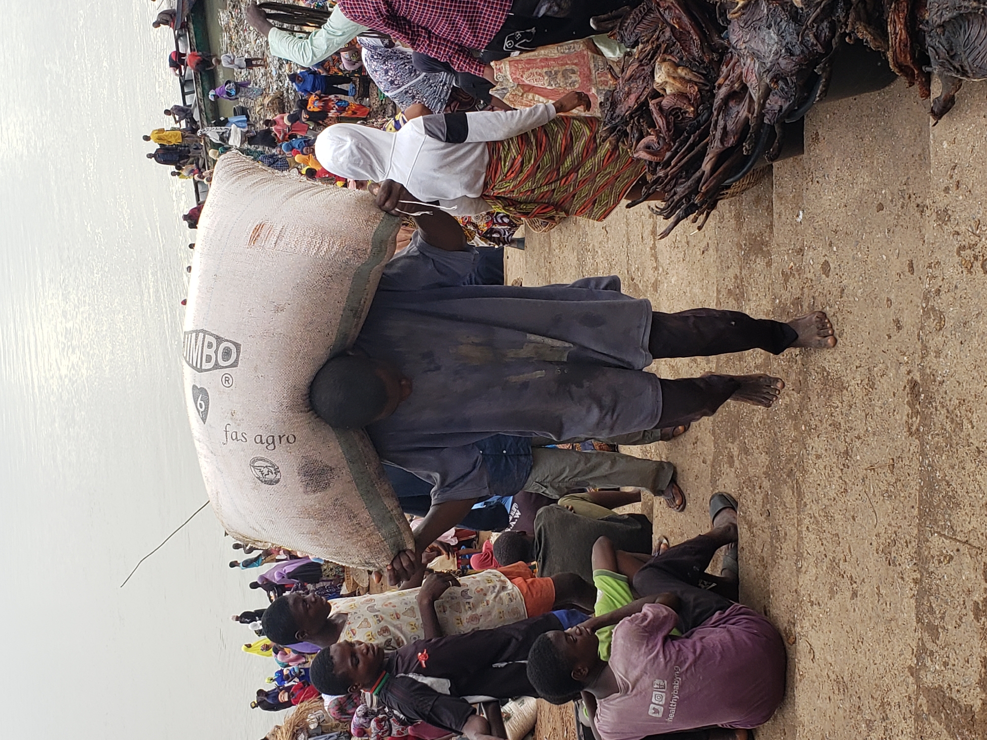 The image shows a man carrying a large sack labeled "JUMBO fas agro" on his back in Lokoja, Nigeria. He is barefoot and wearing dark clothing. Several other people are visible, some lying or sitting on the ground, others standing nearby. One woman wears a white head covering and a patterned skirt. A pile of what appears to be dried meat or fish is to the right. In the background, more people are gathered near what appears to be a body of water. The ground is concrete. The scene suggests a crowded marketplace or port area.