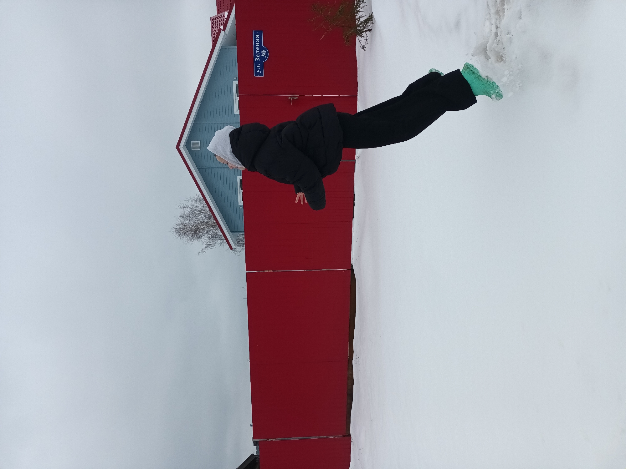 A young person in a black coat and black pants is posed in front of a red building with a blue upper section, standing in snow in Ольховка, Russia. The person is wearing light green boots and has a hooded sweatshirt on underneath the coat. A street sign on the red building reads "ул. Зеленая 30". The sky is overcast. A bare tree branch is visible to the left.