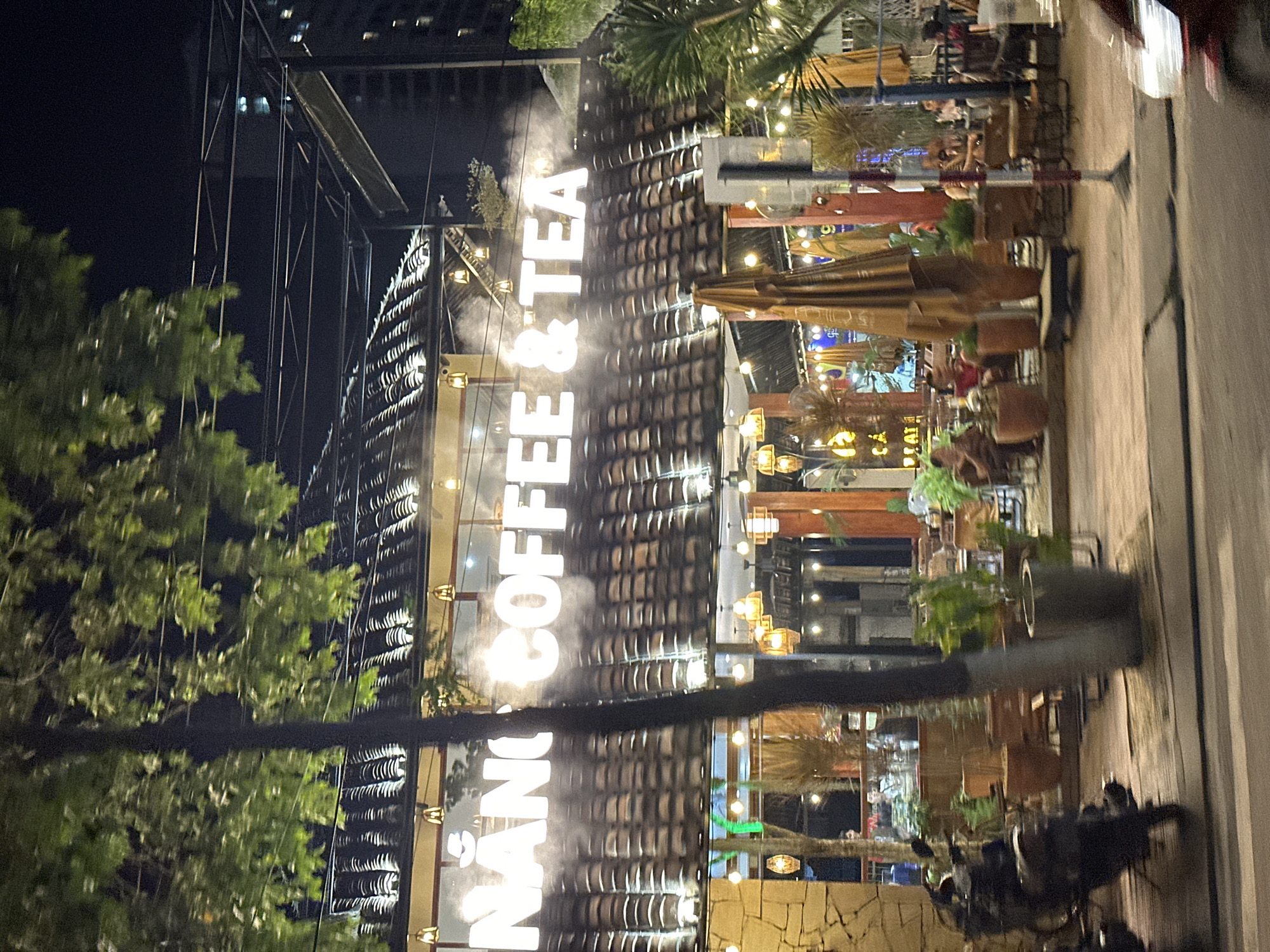 The image captures the exterior of a coffee shop named "Nẵng Coffee & Tea" at night. The coffee shop is located on a street in Bac Nha Trang Ward, Vietnam. People are seated at tables on the patio. The building's facade features a vertical sign with the shop's name. There is decorative lighting. Green trees are visible. A motorcycle is parked nearby. There are buildings and structures in the background.