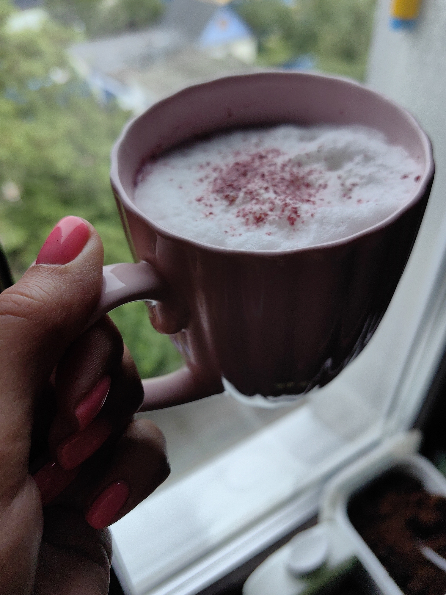 A close-up shot captures a person's hand with bright pink fingernails holding a pink mug filled with a foamy, white beverage dusted with small pink particles. The mug is positioned near a window, with a blurred background showing greenery and hints of buildings, suggesting an urban or suburban setting, possibly in Fanipal, Belarus, given the context. The focus is on the beverage and the hand, with the surroundings providing ambient context.