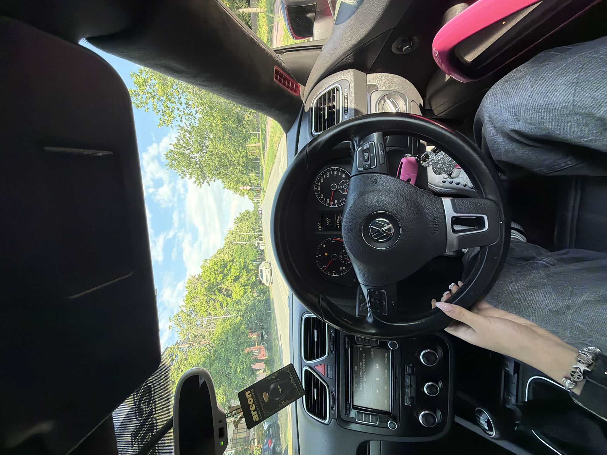 The image is taken from the interior of a car, looking forward. The car is on a paved road with trees lining both sides and a cloudy blue sky above. In the distance, there are a few other vehicles. The dashboard of the car is visible, including the steering wheel, instrument cluster, and a central console with a radio and climate controls. A hand with manicured nails is resting on the steering wheel. A car air freshener hangs from the rearview mirror. A pink item, possibly a bag or accessory, is visible on the passenger seat. The scene appears to be in a suburban or semi-rural area. The time displayed on the car's dashboard is 13:05. The overall impression is a casual snapshot of a person driving on a sunny day.