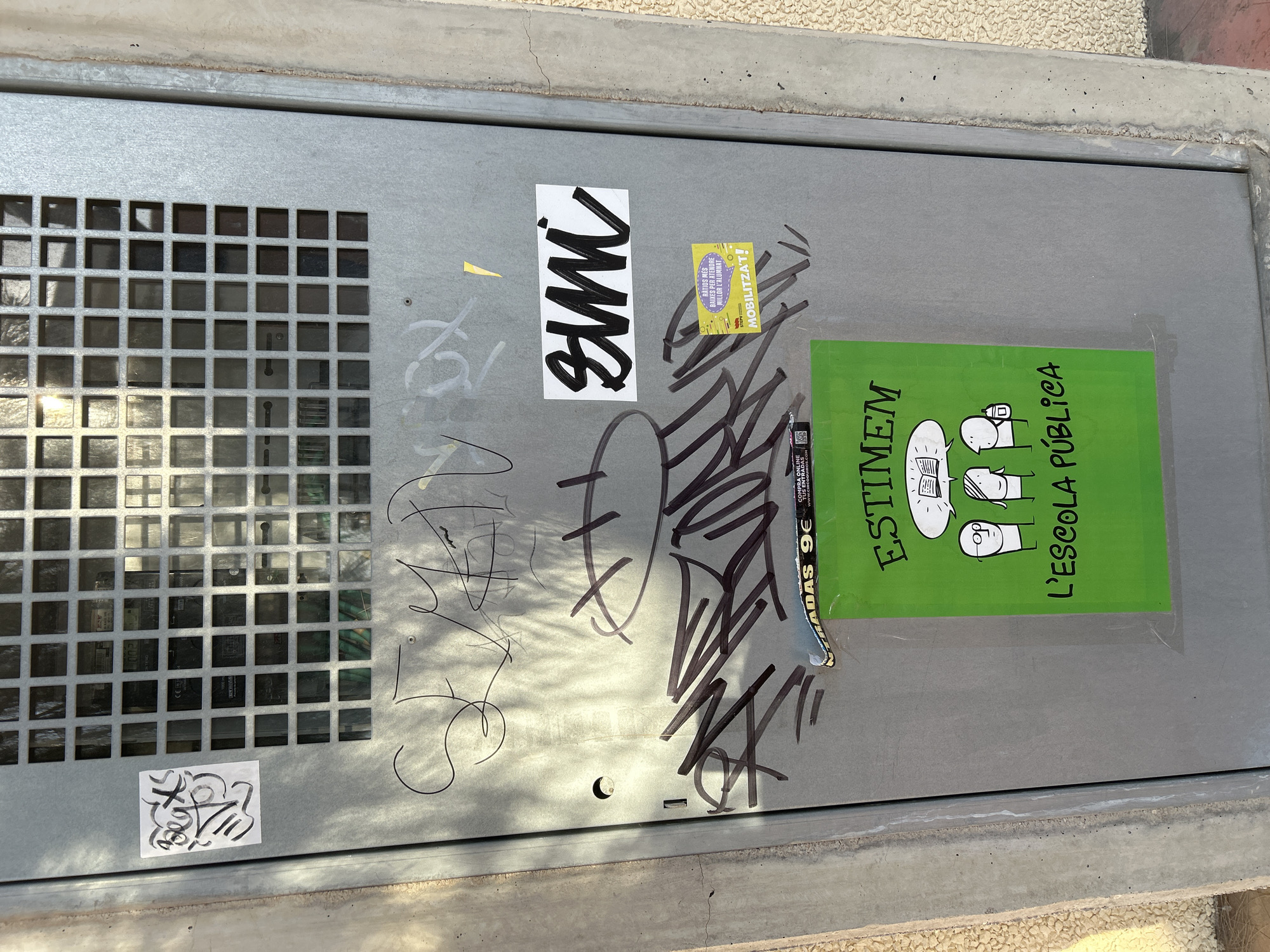 The image depicts a section of a metal utility box on a street in Grao, Spain. The box is covered in various graffiti tags and stickers. A prominent sticker on the lower half of the box is a bright green poster with a cartoon illustration of two figures and text. The poster reads "ESTIMEM L'ESCOLA PÚBLICA" which translates to "We love public school". Above this, there's a sticker with the word "SMI" in bold black lettering on a white background. To the right of the "SMI" sticker, a smaller yellow sticker with Catalan text, "RATIOS RES BAIXES PER ATENDRE MILLOR L'ALUMNAT MOBILITZA'T!", meaning "Fewer low ratios to better serve the students, mobilize!". There is also a torn sticker above these that reads "ENTRADAS 9€". The metal box has a grid of square openings at the top, revealing some inner components. The surrounding environment includes concrete elements and a textured wall, typical of urban street furniture. The lighting suggests it is daytime, with shadows cast across the surface.