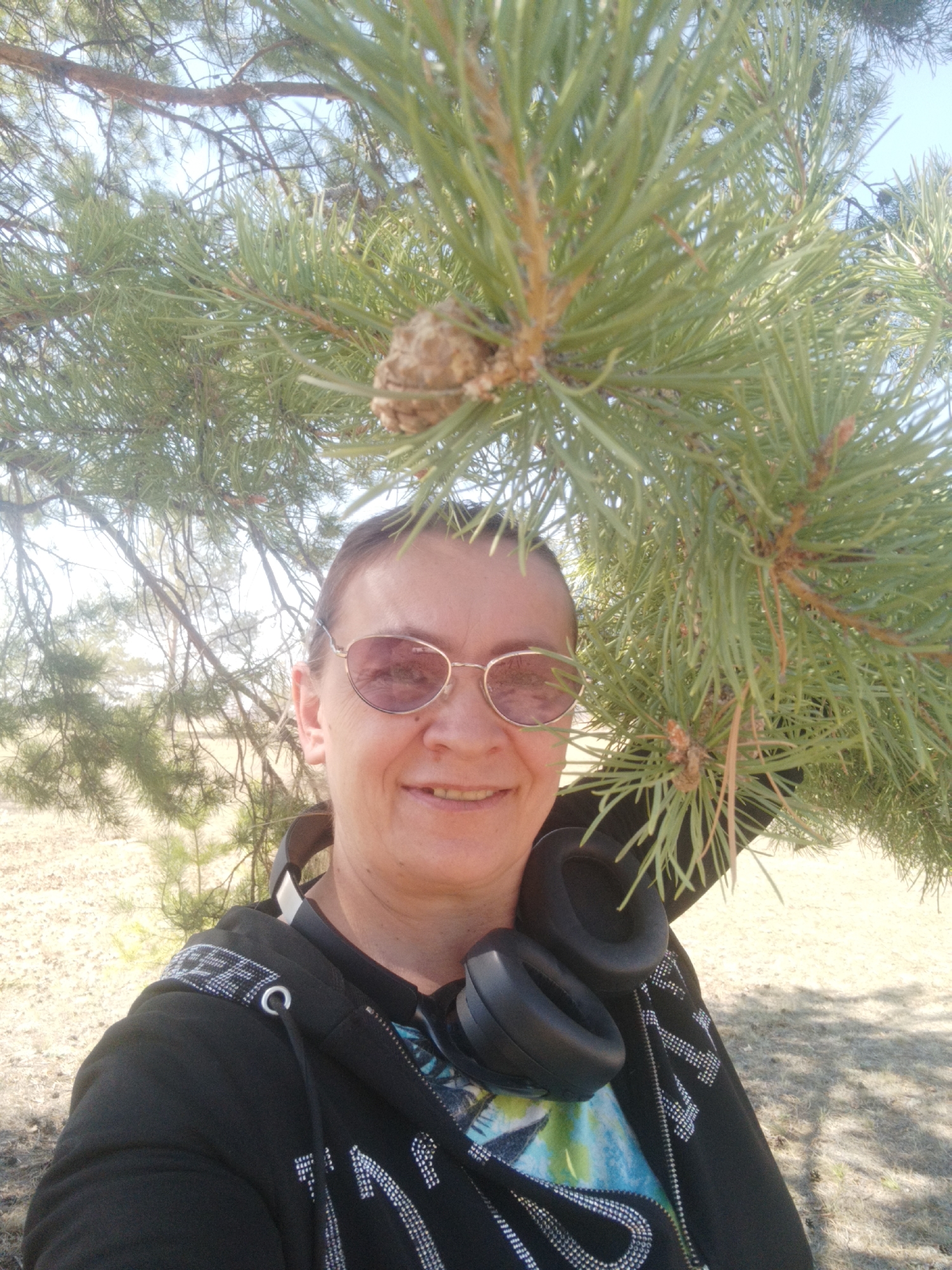 A middle-aged woman is posing for a photo outdoors. She has short brown hair, is wearing rose-tinted glasses, and a black hoodie with white embellishments. Around her neck, she has a pair of black headphones. She is smiling. The background shows a dry, grassy area and trees. Coniferous branches with a pinecone are prominently featured in the foreground, partially obscuring the top part of the frame. The location is identified as Syrdakh, Russia.