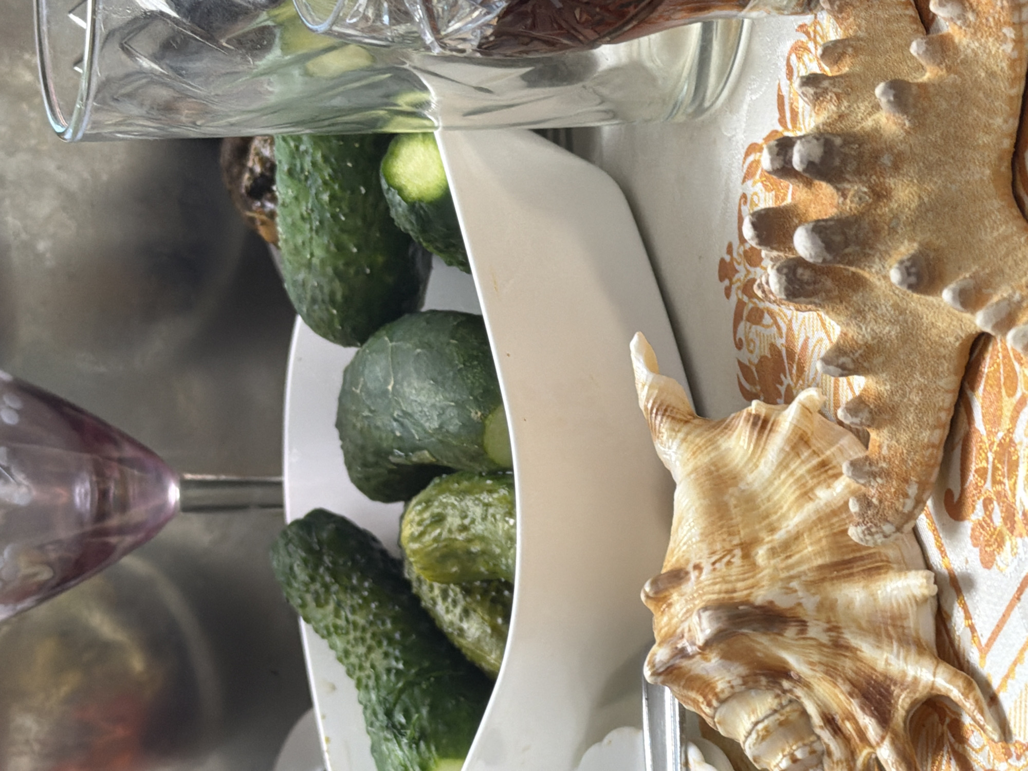 A still life composition features a white bowl filled with pickles, a seashell, and a starfish arranged on a patterned tablecloth. In the background, a glass and a stemmed glass with a purple hue are visible. The scene is set indoors, possibly on a table, and does not provide any specific clues to its location within the city of Bronnitsy, Russia. There are no people or actions depicted.