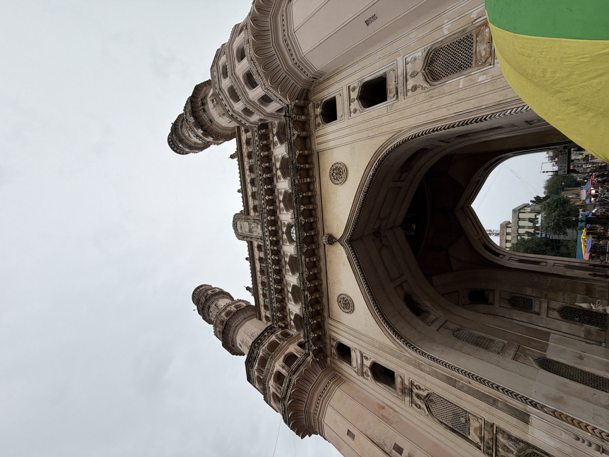 The media file shows a low-angle shot of the Charminar monument in Hyderabad, India, against an overcast sky. The intricate architectural details of the monument, including arches, minarets, and decorative carvings, are prominently featured. The texture and material of the monument appear to be light-colored stone, possibly sandstone.
The scene is set in an outdoor environment, suggesting a public space around the monument. The lighting indicates it might be daytime, but the cloudy sky suggests a lack of direct sunlight, perhaps a subdued or gloomy atmosphere. Some people and activity are visible in the background, seen through the archway of the Charminar, hinting at a busy area with ongoing public life, possibly a market or street scene. A large, brightly colored fabric, split between yellow and green, is draped over the right side of the frame, partially obscuring the monument and adding a splash of color to the otherwise muted tones of the stone and sky. This fabric could be part of a stall, a banner, or a decorative element.
The overall impression is of a monumental structure in a lively urban setting, captured from an unusual perspective that emphasizes the grandeur and complexity of its design. The image focuses on the architectural elements, with the sky and a large piece of fabric dominating the composition.