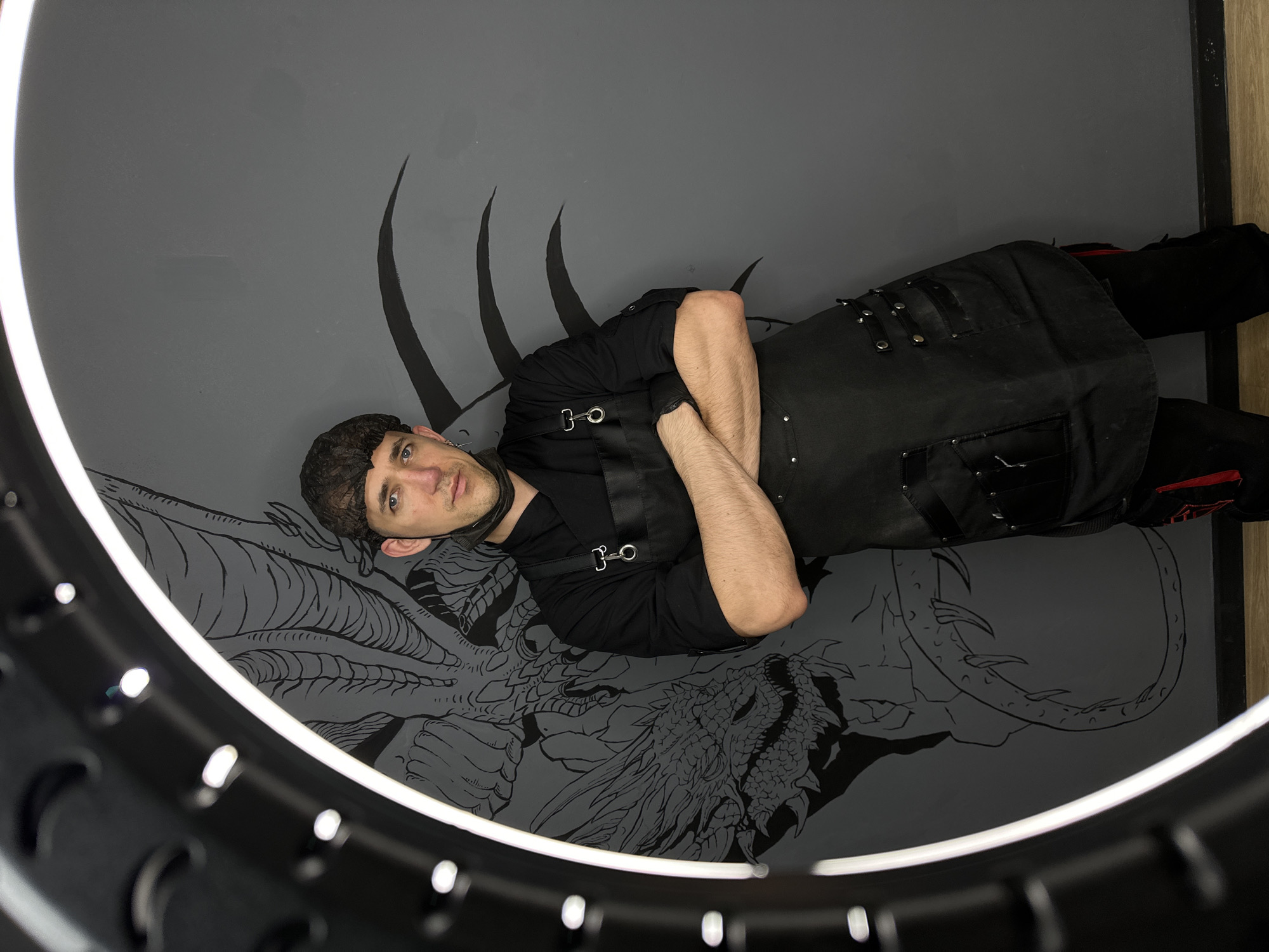 In Saratov, Russia, an individual stands against a gray wall decorated with a black dragon mural. The person, who appears to be male, is wearing a black chef's coat, a black apron, and dark pants with red accents. A dark hairnet covers their hair, and a black face mask is pulled down to their chin. Their arms are crossed, and they are looking directly at the camera. In the foreground, a circular light fixture with multiple bulbs frames the scene. The lighting is bright, casting some reflections on the glossy surfaces. The floor is made of light-colored wood.