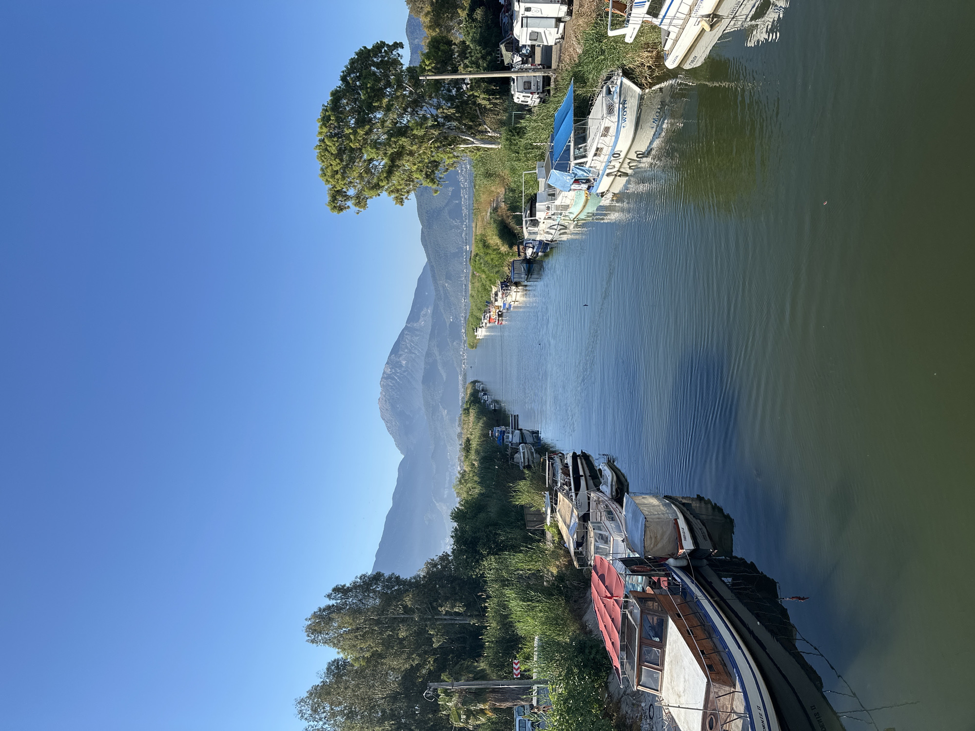 The image depicts a riverbank scene in Fethiye, Turkey. Along a stretch of the river, numerous boats of varying sizes and conditions are moored, some appearing older and possibly houseboats. Lush green vegetation lines both sides of the river, with trees interspersed among the boats. In the background, a mountain range rises, partially shrouded in a haze. The sky is clear and blue, indicating a sunny day. Some vehicles, possibly RVs or campers, are parked near the boats on one side of the river.