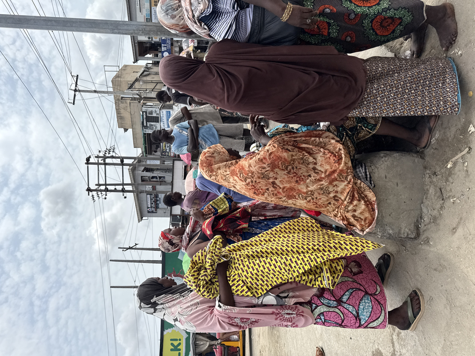 This media file captures a bustling street scene in Lagos, Nigeria, during daylight hours under an overcast sky. The image is oriented vertically (sideways), showing a crowd of people, predominantly women, gathered along what appears to be a sidewalk or roadside.
**Subjects:** The foreground features several women dressed in vibrant, patterned traditional West African attire, including headscarves and flowing garments. Colors like yellow, pink, orange, blue, and brown dominate their clothing. Some women are seated on low concrete ledges, while others stand. Many are wearing sandals or are barefoot. In the background, several men are also visible, one in a light blue t-shirt and another in a collared shirt or suit. Urban buildings, possibly shops or residences, line the street. Prominent utility poles with numerous overhead power lines crisscross the sky, a common sight in Nigerian cities.
**Activity or event taking place:** The scene suggests an ordinary daily gathering, possibly at a market, bus stop, or a busy commercial area. People appear to be waiting, observing, or engaging in various everyday activities. The atmosphere is generally calm, with no strong emotions discernible on faces.
**Notable details:** The diverse and colorful clothing is a significant detail, reflecting local fashion. The infrastructure, with its dense network of power lines, provides a clear geographical cue. The ground is paved, likely concrete.
**Visible text:** Several signs are visible in the background: "solar" and "08" (possibly part of a phone number) are seen on upper signs. Lower down, "JERRYTECH" and "LAPTOPS" are clear on another sign, suggesting electronic sales. A green and white sign with "LK!" is partially visible at the bottom left. "WI" is also visible on a sign.