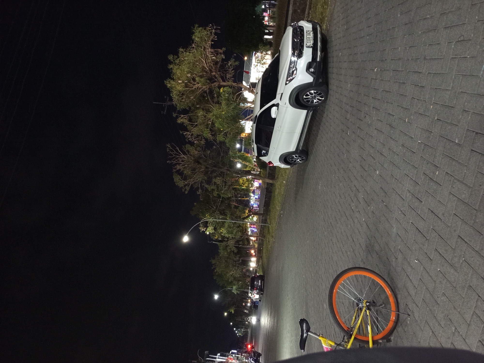 The image depicts a street scene at night in Gemurung, Indonesia. A white SUV is parked on the right side of the paved road. In the foreground on the left, a yellow bicycle with an orange rim is partially visible. The street is lined with trees and illuminated by streetlights, with distant lights suggesting buildings or businesses. The sky is dark.