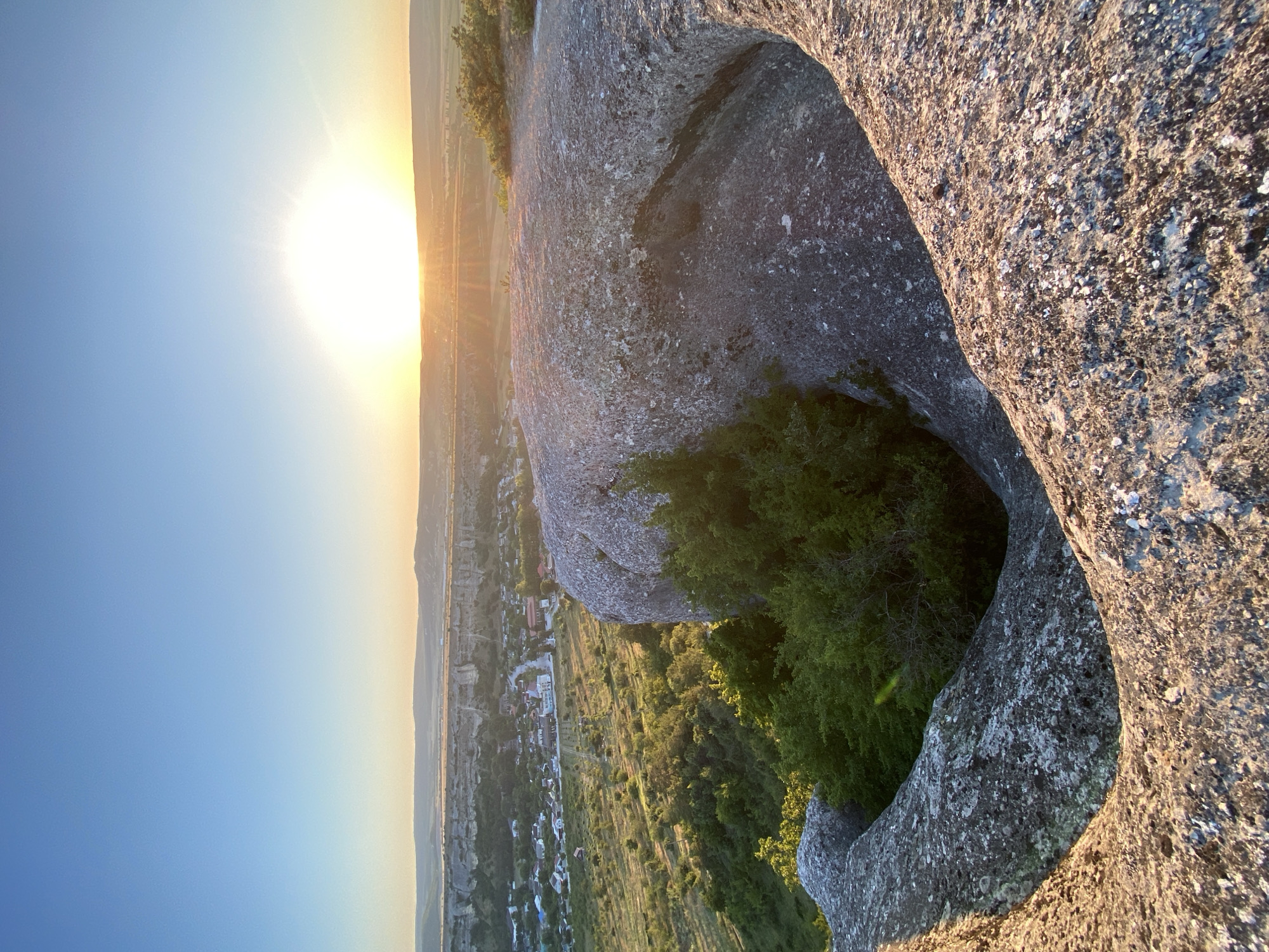 This is a vertical, eye-level shot of a rocky landscape at sunset, likely taken from a high vantage point. In the foreground, large, weathered rocks dominate the frame, with a natural opening or hollow in the center of the right-hand rock. The rocks are rough and textured, with patches of lichen and moss.
Below and in the middle ground, a small village is nestled in a valley. The houses have red or brown roofs, and a road winds through the settlement. The surrounding landscape is a mix of green fields, rolling hills, and dense patches of trees. In the distance, a long, flat-topped mesa or plateau stretches across the horizon.
The sky is clear and blue, transitioning to a warm orange and yellow near the horizon where the sun is setting. The sun itself is a bright, intense orb with visible rays fanning out. The overall impression is one of a serene and expansive natural landscape bathed in the warm light of a setting sun, with a small human settlement integrated into the environment. The specific location is within the Krasnomakskoye rural settlement in Ukraine.