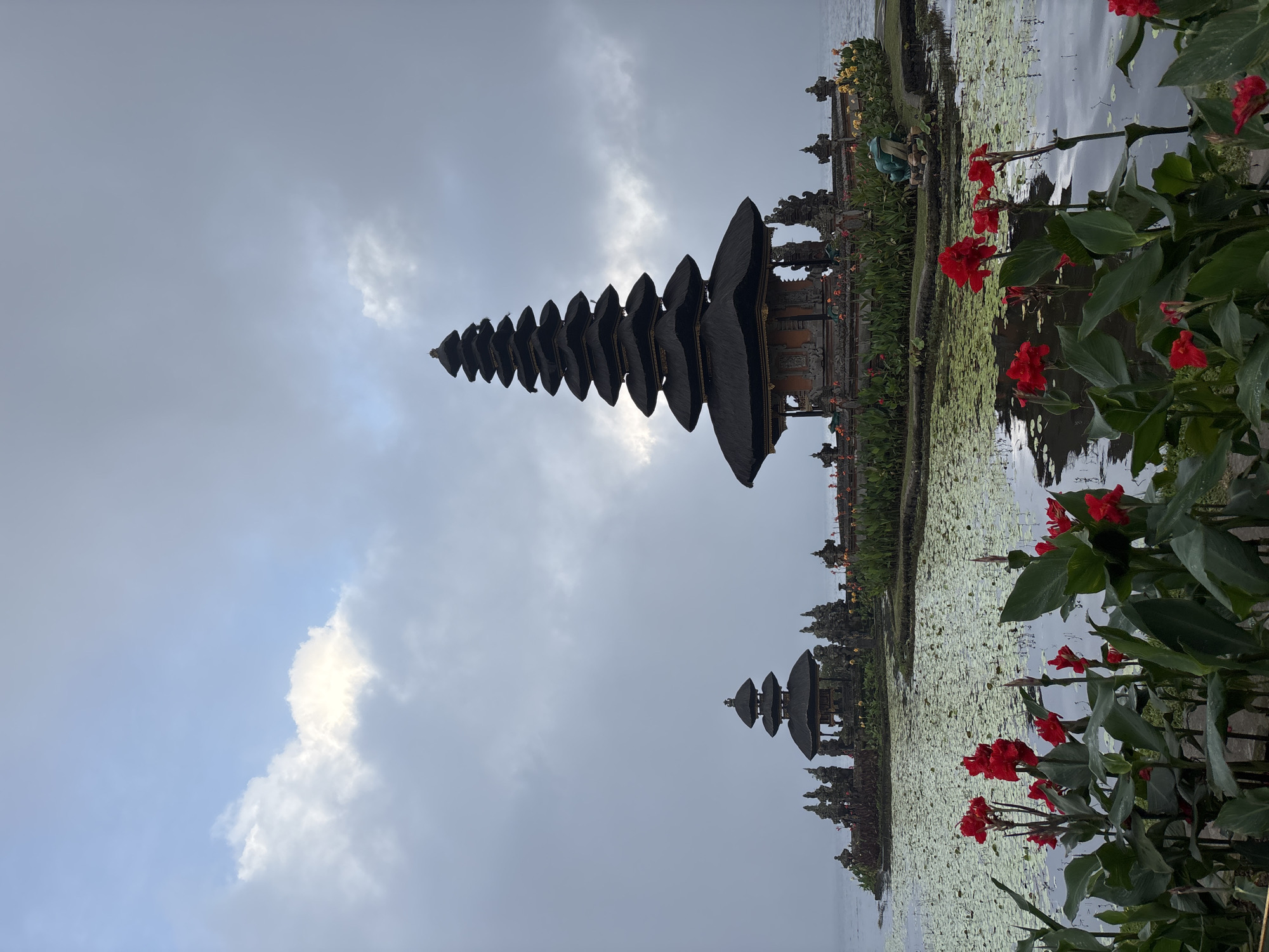 The image depicts the Ulun Danu Beratan Temple in Baturiti, Indonesia. The temple is a major landmark, situated on the edge of Lake Bratan. The distinctive multi-tiered Meru towers of the temple complex are reflected in the calm water of the lake. Red flowers with green leaves are visible in the foreground on the right side of the image. The sky is overcast, with a mix of clouds and some patches of light blue visible. The image is oriented vertically, resulting in an inverted view of the scene.