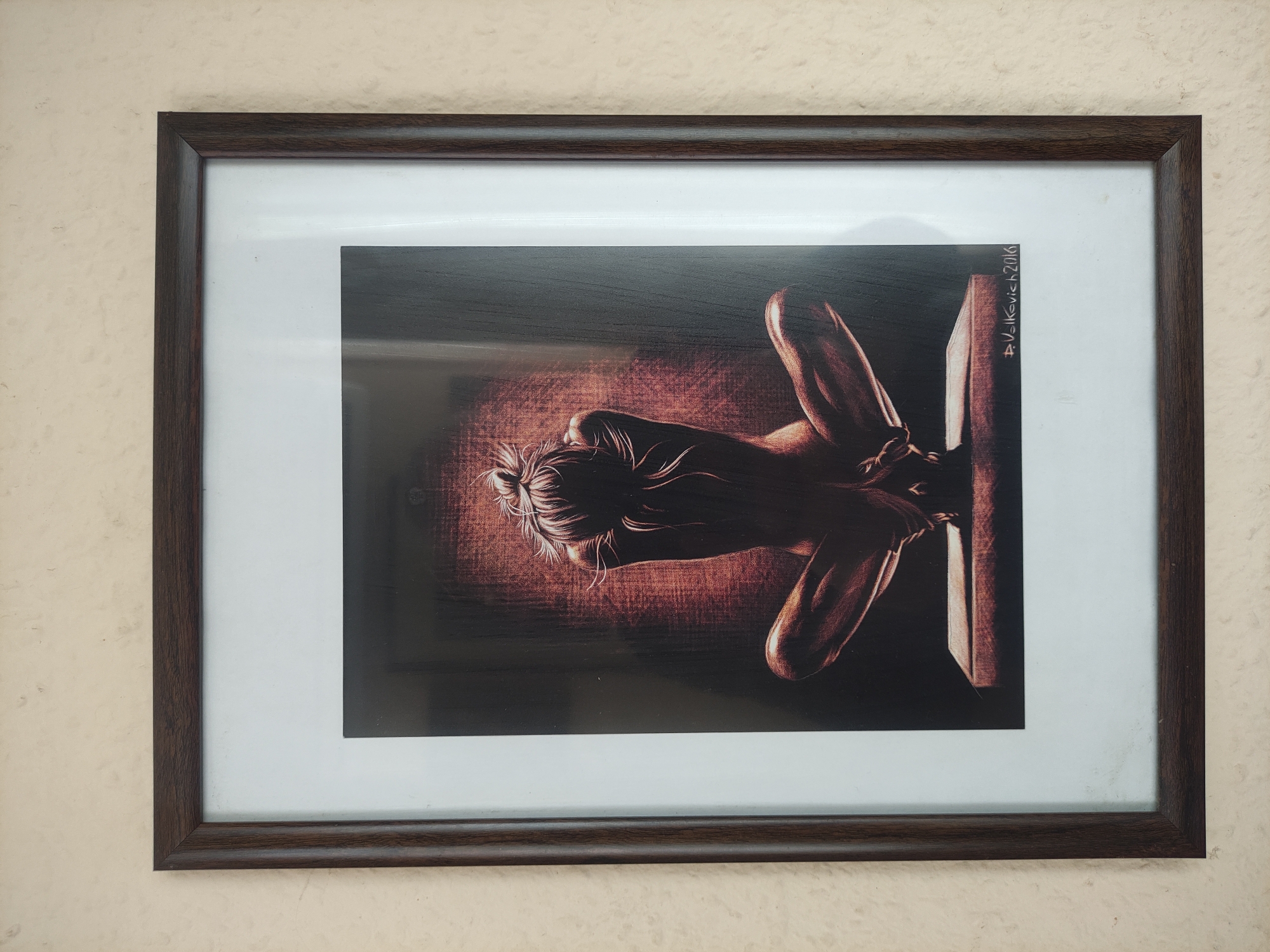 A framed photograph hangs on a light beige wall. The frame is dark brown. Inside the frame is a print featuring a monochromatic image of a woman with her hair in a bun. She appears to be bound to a wooden structure. The print bears the signature "A. Volkevich 2016" in the upper right corner. The location is unknown, but the city is Tbilisi, Georgia.