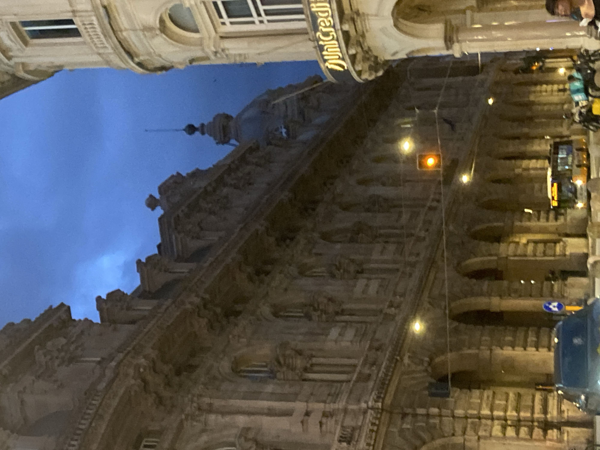 The image captures a street scene in Genoa, Italy, at dusk. A grand, ornate building with arched windows and detailed architectural features dominates the left side of the frame. On the right, another building, lighter in color and with a curved facade, features a sign that reads "UniCredit." The sky above is a deep blue, indicating twilight. Several streetlights are illuminated, casting a warm glow on the buildings and street. In the foreground, a bus is visible, along with several motorcycles and a car. A pedestrian is also present on the right side of the image. The overall impression is that of a bustling urban environment transitioning into the evening.