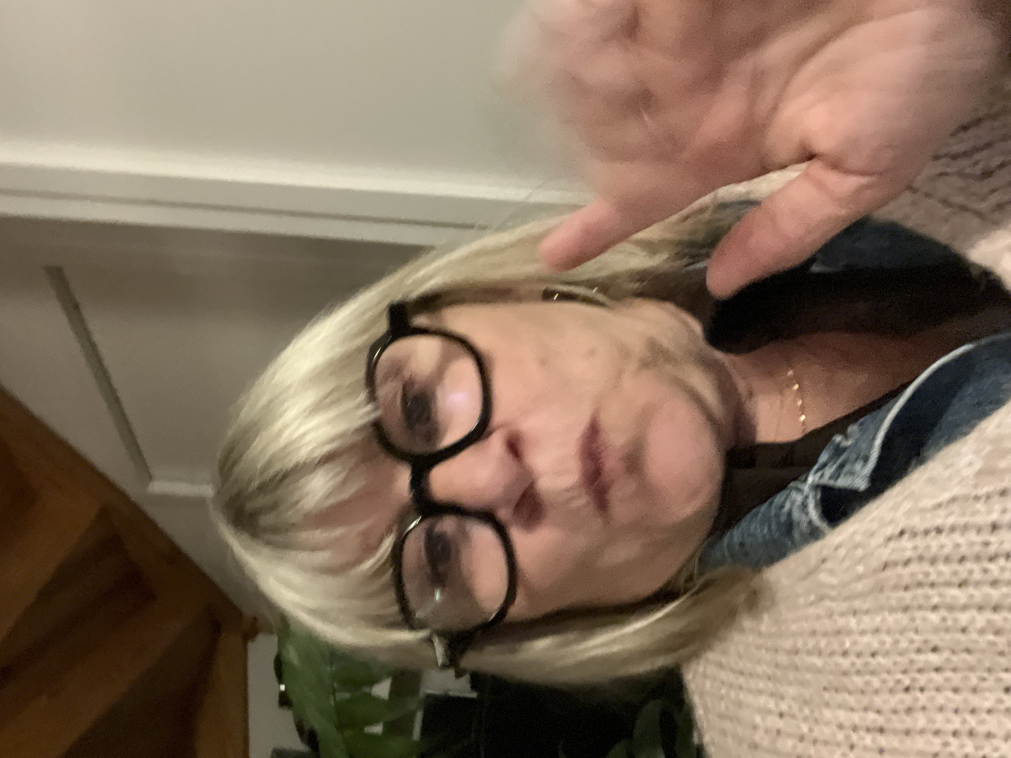 An indoor portrait image, captured in Chambœuf, France, features a Caucasian woman with blonde hair and bangs, wearing prominent black-framed glasses. She is positioned centrally, her face exhibiting a puckered expression with slightly parted lips. Her attire includes a light-colored, chunky knit sweater over a dark garment, possibly a denim shirt, and a delicate gold-toned necklace is visible around her neck. Her right hand (viewer's left) is raised and exhibits motion blur, extending towards the right side of the frame. The background to the left reveals dark green foliage from a plant and angled dark wooden ceiling beams. The background to the right consists of a light-colored wall with a distinct vertical architectural element, likely a door frame. The scene is illuminated by artificial light.