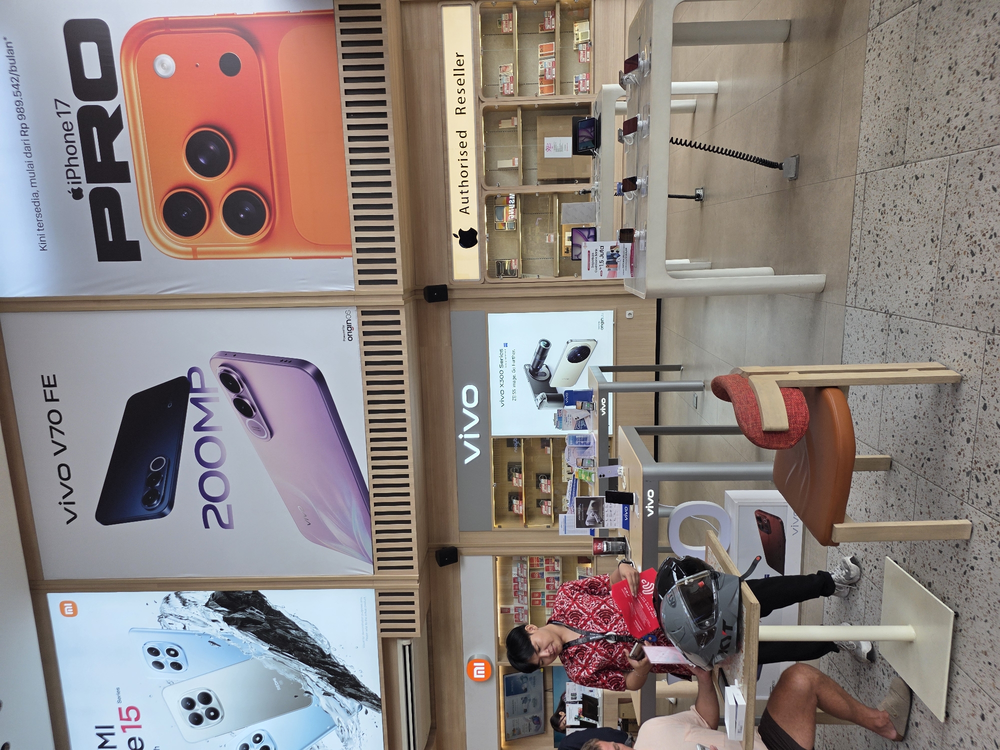 Here is a forensic description of the scene:
The image appears to be taken inside a mobile phone store in Unknown, Unknown. The scene is well-lit and features several large advertising displays for various phone brands and models. Visible brands include iPhone, Vivo, and Redmi.
The advertising displays feature images of the phones, the phone names, model names, and some pricing information. In particular, an orange iPhone 17 PRO, a purple Vivo V70 FE featuring "200MP", and a Redmi series phone are prominently displayed.
Along the back walls of the store are displays containing phones for sale. There are customers inside the store, and at least one employee is present. One customer is seated at a table and appears to be examining information, while a helmet sits nearby. Another customer is also seen seated.
The store also has a section labeled as "Authorised Reseller" (likely for Apple products), with a corresponding Apple logo. The floor is tiled with a speckled pattern.