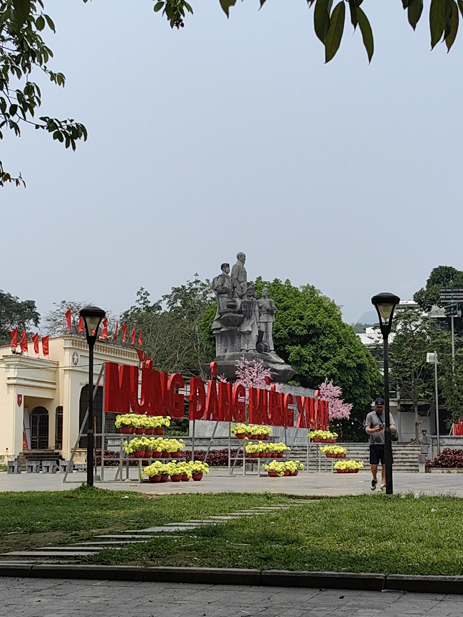 The image shows a public square in Thành phố Hà Giang, Vietnam. In the foreground, there is a patch of green grass with stone pavers leading toward a paved area. In the middle ground stands a large red sign with Vietnamese characters, adorned with pots of yellow flowers. Behind the sign, there is a statue of several figures. To the left, a building with red flags is visible. A man is walking on the right side of the square. Tall trees surround the area. The sky is overcast. The light posts are visible on both sides of the image.