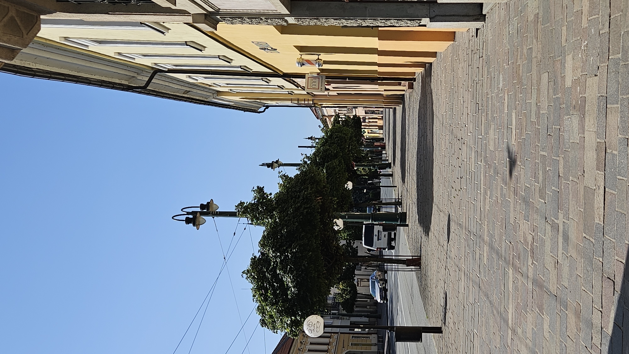 The image depicts a street scene in Prešov, Slovakia, likely on the main pedestrian zone or a similar prominent thoroughfare. On the right side of the frame, a row of colorful buildings lines the street, with facades painted in shades of yellow and orange. One building features a barbershop pole, indicating a commercial area. A sidewalk paved with stone slabs runs alongside the buildings, leading into the distance.
On the left side of the image, a street with light traffic is visible, including a silver car and a white van. A row of trees with lush green foliage is planted along the sidewalk, providing shade. Several ornate streetlights with double lamps are spaced along the pedestrian walkway. In the foreground, the paving stones of the sidewalk are clearly visible, with shadows cast by the trees and streetlights. The sky is a clear, bright blue, suggesting a sunny day. The scene is tranquil and appears to be captured in the daytime.