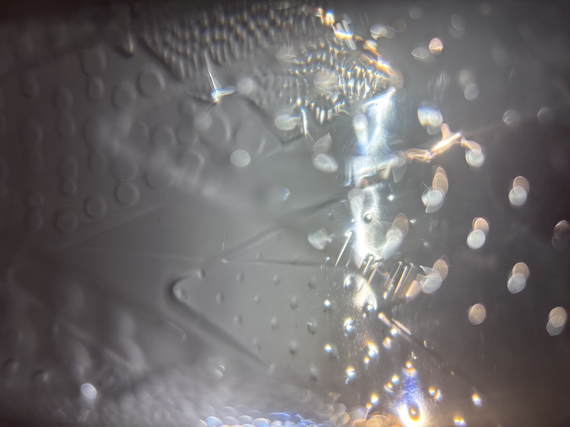 The image is a close-up, abstract shot of water droplets on a textured surface. The water creates a refractive and distorting effect on the surface beneath, which appears to have raised circular patterns and wavy lines. Bright highlights and bokeh effects are prominent due to the lighting interacting with the water and surface. The overall color palette is muted grays, whites, and subtle iridescent tones from the light reflections. No specific location within a city can be determined.