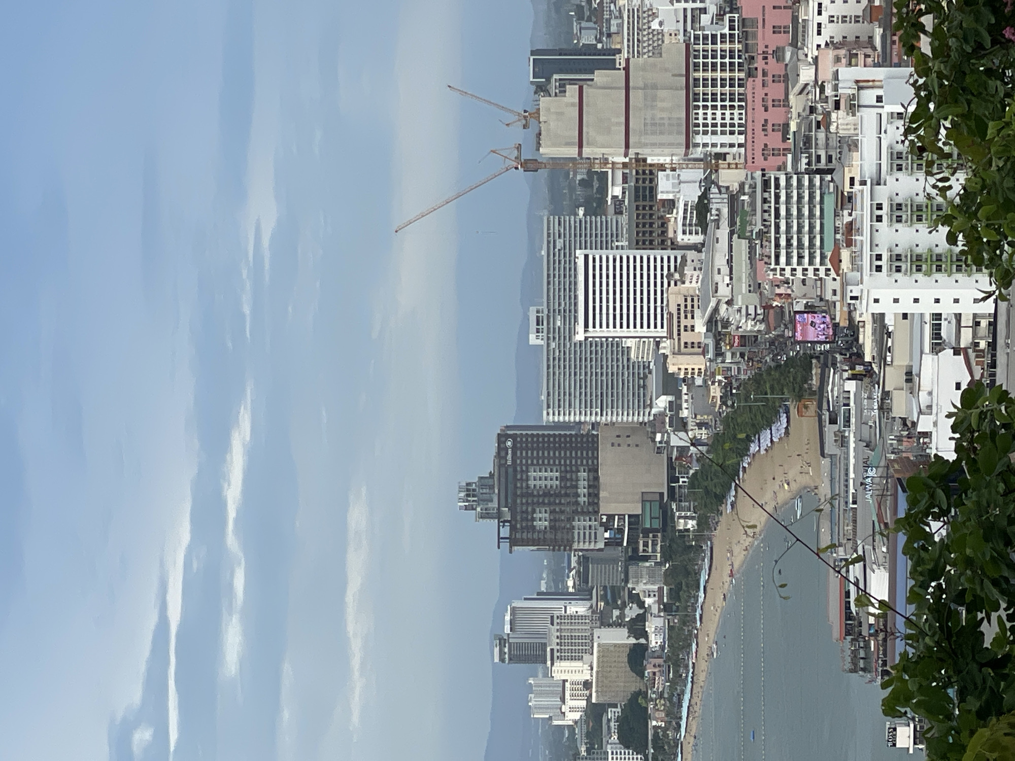 An elevated, wide shot captures a panoramic view of Pattaya City, Thailand, on an overcast day. The cityscape is dominated by numerous high-rise buildings of varying architectural styles and colors, including hotels, apartments, and office buildings. Two large construction cranes are visible, indicating ongoing development. In the foreground, lush green foliage partially obscures the lower portion of the image. A busy beach stretches along the coastline, dotted with umbrellas and filled with people enjoying the sand and sea. Boats are scattered on the water, and a road with some traffic runs parallel to the beach. The ocean meets the sandy shore, and a hazy mountain range can be seen in the distance, silhouetted against the muted sky.