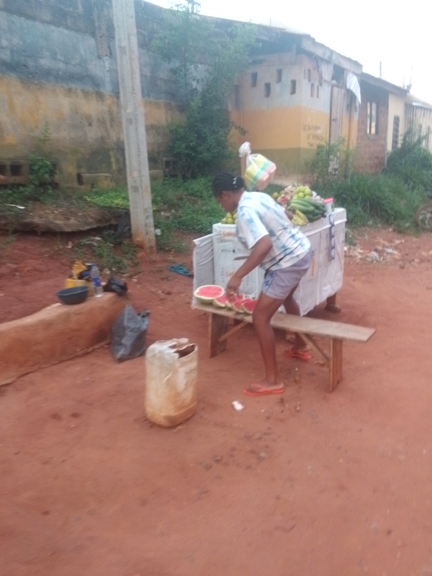 In Nkpor, Nigeria, a woman is standing at a table beside the road selling fruit, including watermelon, which she is cutting into slices. The area is dusty red clay. Other items and objects are near the table, including a water jug and a bench. A concrete wall is visible in the background behind a utility pole. Across the road, a building with a yellow and white exterior stands partially hidden by foliage.