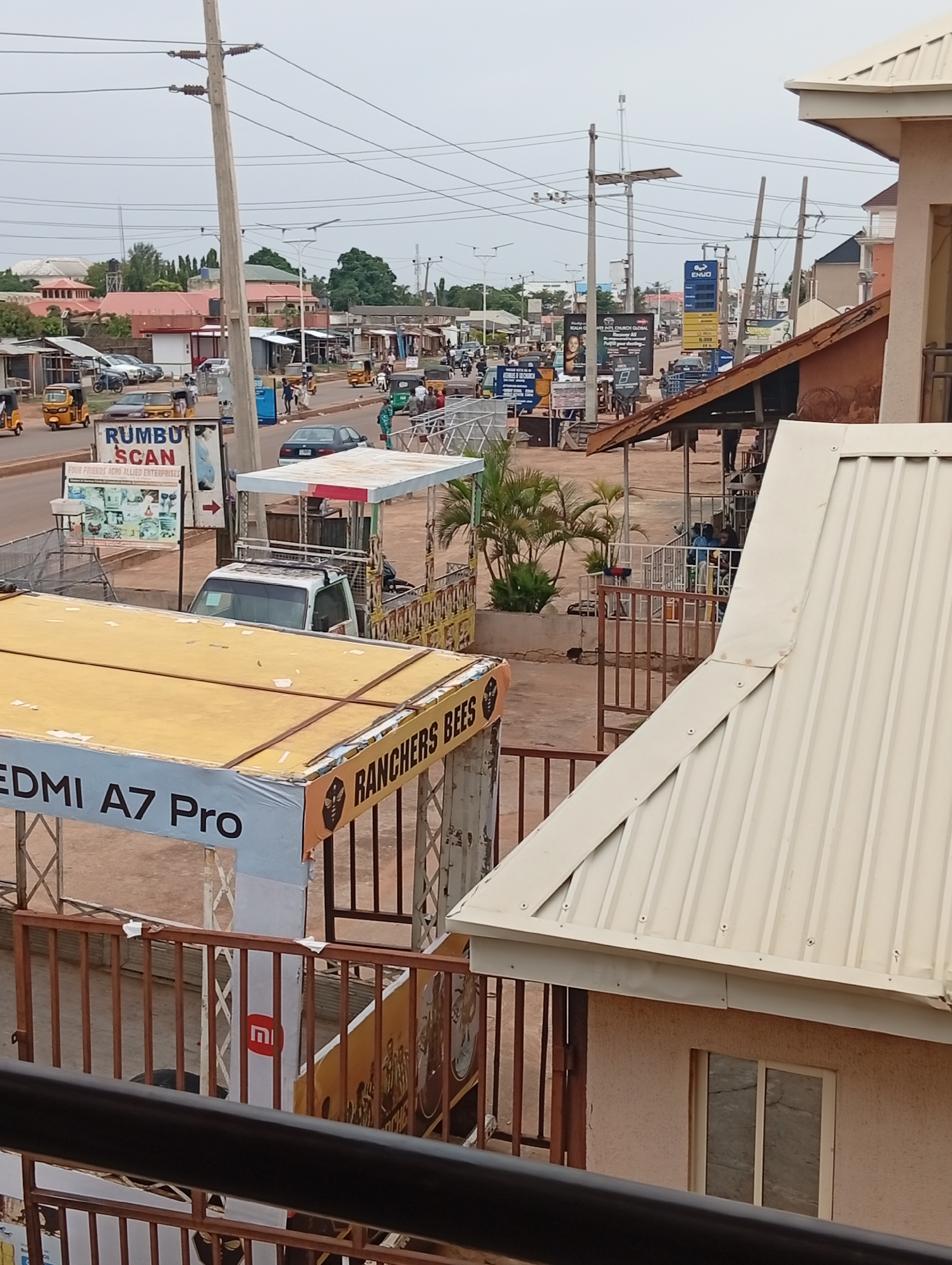 The image shows a street scene in Sabon Tasha, Nigeria. A street with power lines overhead is visible in the background, populated with people, vehicles, and small shops. Prominent signage for "RUMBU SCAN" and "RANCHERS BEES," as well as promotional materials for "REDMI A7 Pro," are situated in the foreground. Various pedestrians and commercial tricycles (keke) are visible along the street.
