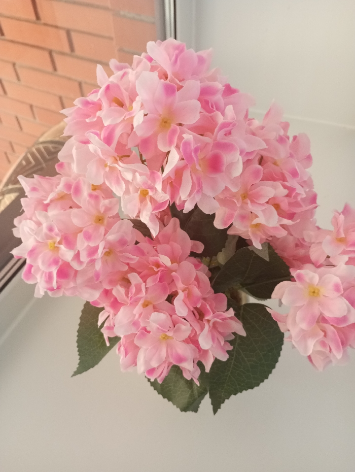 A close-up forensic examination of the scene in Yaromaska, Russia, reveals a large bouquet of artificial pink flowers, likely hydrangeas, centrally positioned. The flowers consist of numerous small, light pink petals with some exhibiting darker pink edges, and faint yellow centers. Dark green artificial leaves are visible underneath and between the flower clusters. The objects are situated indoors, near a window. Through the window on the left, a reddish-brown brick wall with light-colored mortar lines is visible, along with a partial view of a dark, patterned object. A vertical, light-colored window frame separates the outdoor view from an indistinct, plain white or light-colored wall or surface that forms the background to the right and lower portion of the image. No people are present, and no active interactions or actions are recorded.
