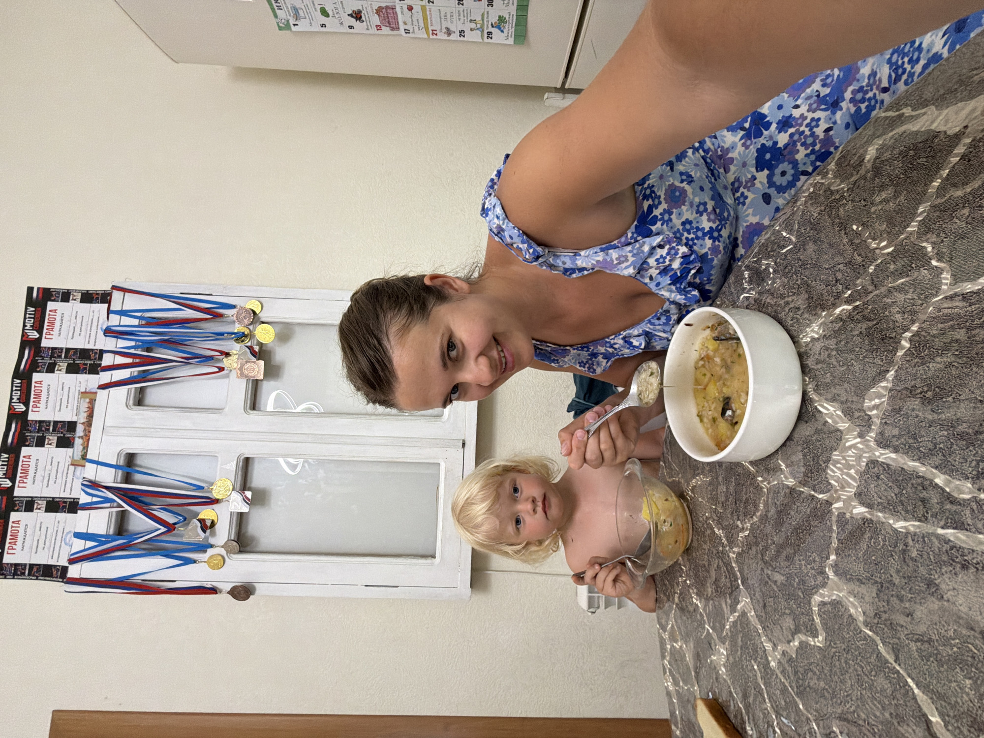 A woman and a young child are seated at a marble-patterned table, about to eat soup. The woman, in a blue floral dress, is holding a spoon with soup, looking towards the camera with a smile. The child, a blonde boy, is shirtless and has a bowl of soup in front of him, also holding a spoon. Behind them, a window with white shutters is adorned with several medals and what appear to be certificates. The setting appears to be a home, possibly in Sochi, Russia, as indicated by the calendars on the right wall. The overall atmosphere is casual and familial.