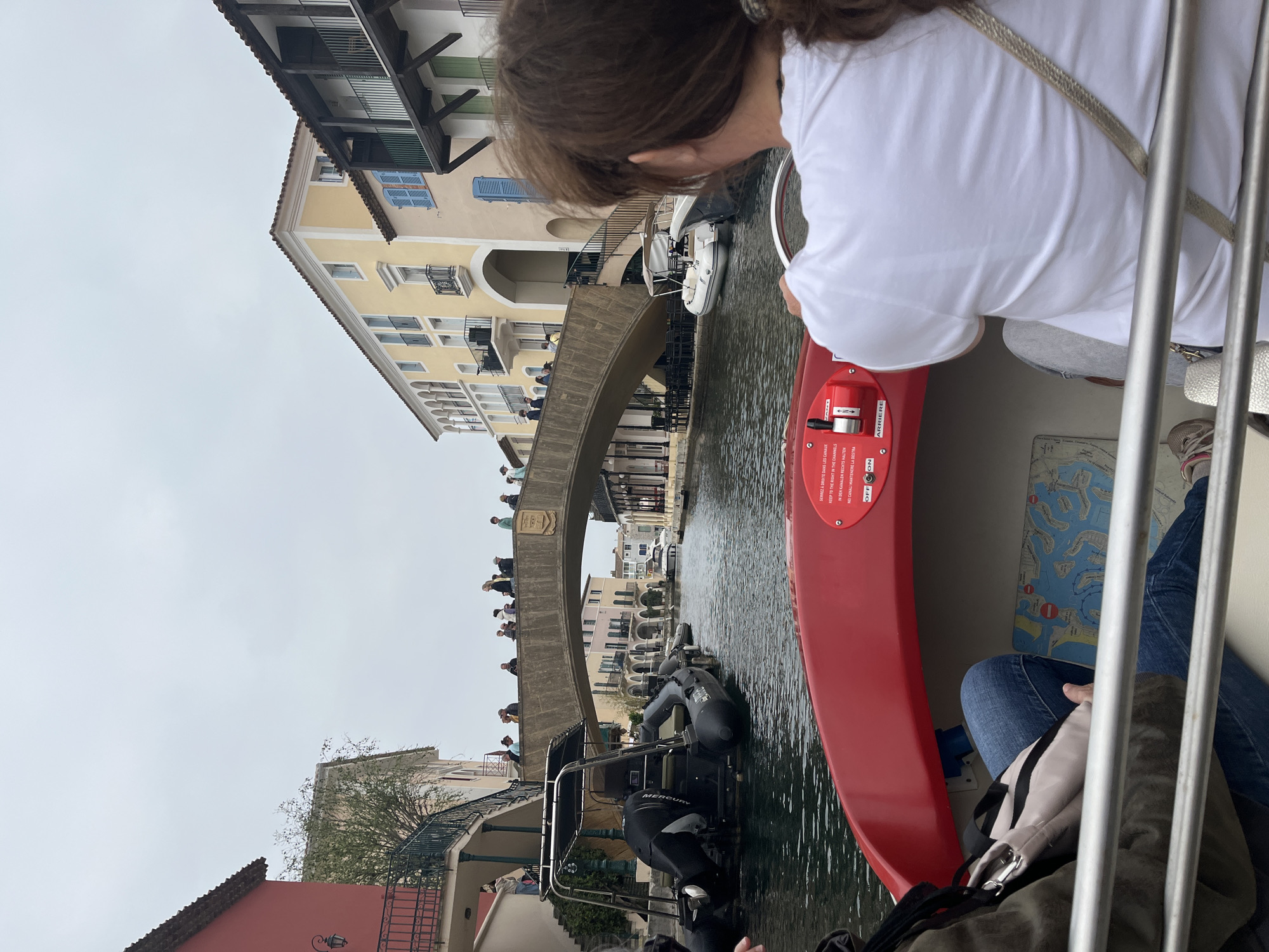 A slightly high-angle, rotated indoor-outdoor shot captures a water-level perspective from a small canal boat in Grimaud, France. A woman with light brown, shoulder-length hair, wearing a white t-shirt and light grey jeans, is facing forward, presumably steering the red-trimmed boat. A second individual in jeans and seated is partially visible in the lower right quadrant.
The boat navigates a canal lined with buildings of varying heights and architectural styles. On the left, a red-roofed building is followed by a taller structure with yellow-beige facades and blue shutters. A stone bridge spans the canal, topped with a crowd of people peering over the edge. Another boat is behind the first boat. The canal is filled with green water. The sky is overcast.