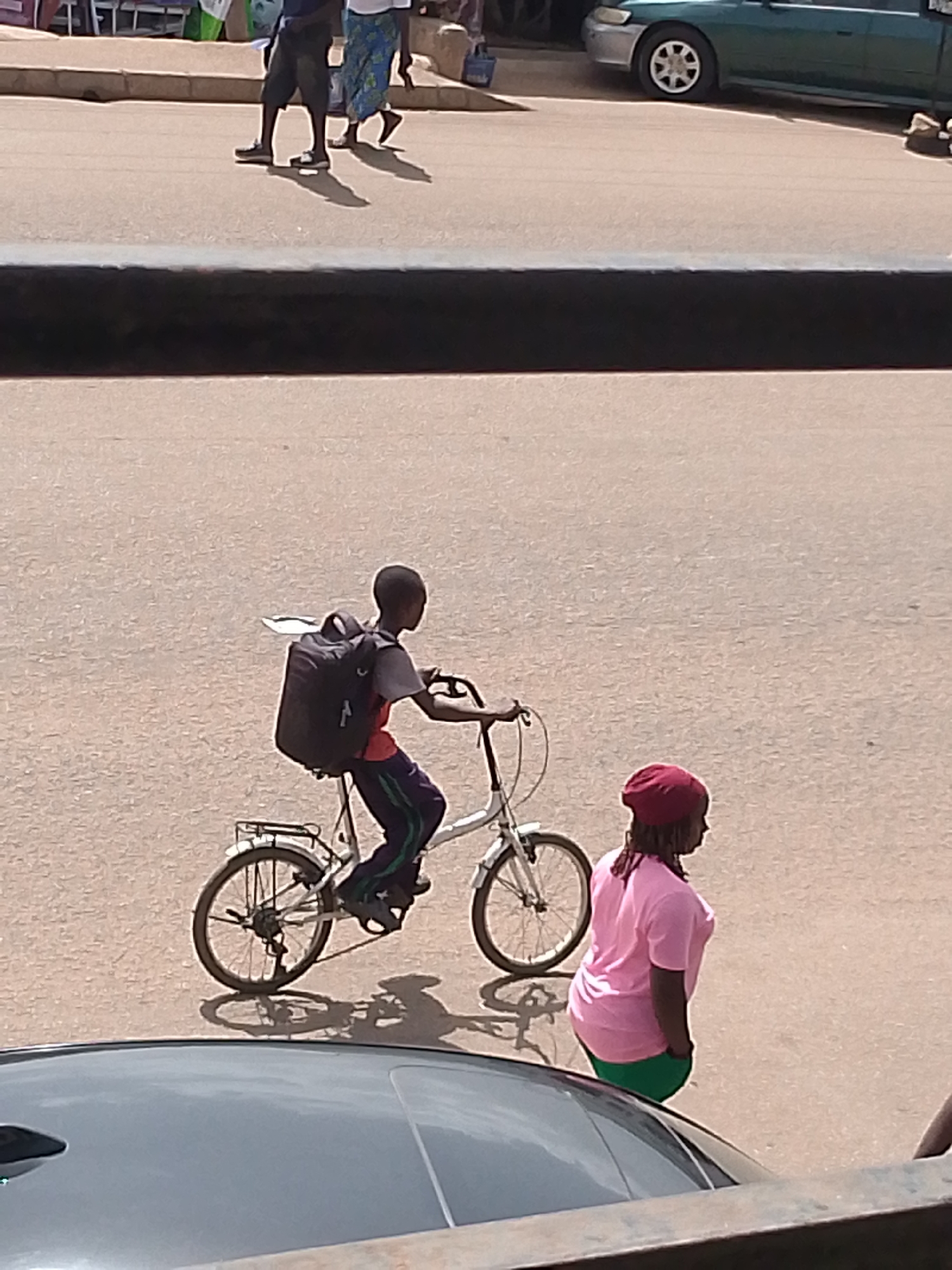 In Bukuru, Nigeria, a boy is riding a bicycle with a large black backpack on his back. He is wearing a gray t-shirt and dark pants with a green stripe down the side. To his right, a woman wearing a pink t-shirt and green pants stands facing away from the camera. They are on a paved street. In the background, a green car is parked. The overall scene depicts everyday life on a street in Bukuru.