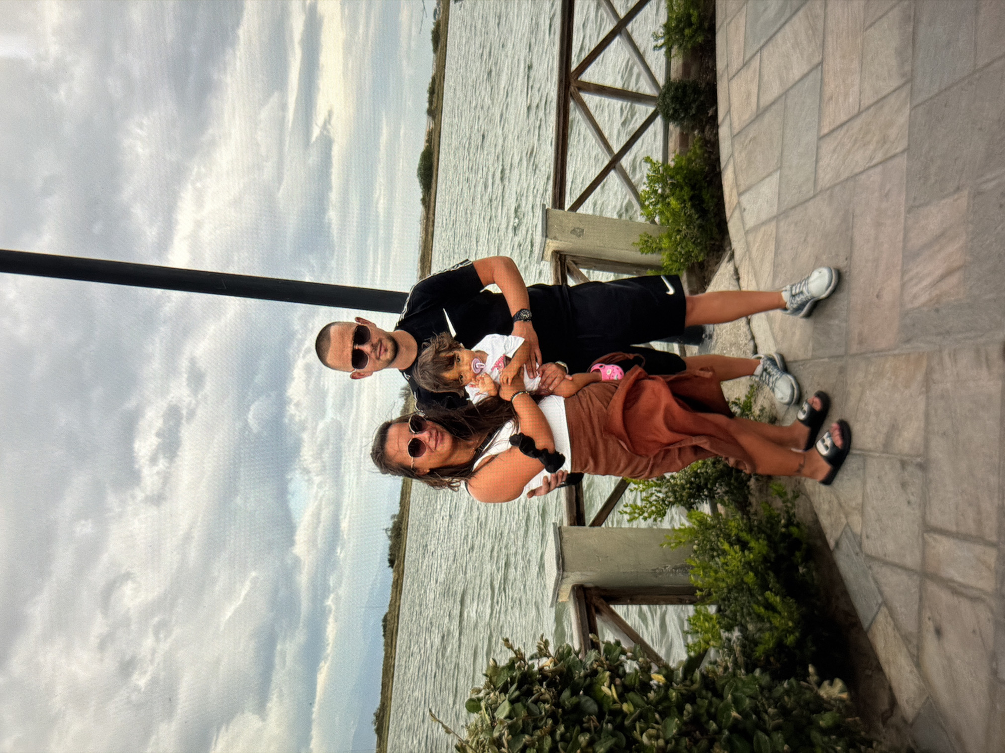 An outdoor, eye-level shot captures a family of three – a man, a woman, and a young child – standing together on a paved walkway next to a body of water under a cloudy sky. The image is oriented sideways, but assuming correct rotation, the man stands on the left, embracing the woman and child who are positioned slightly to his right.
The man, bald and wearing sunglasses, a black t-shirt, and black shorts, smiles faintly. A white Nike swoosh is visible on his shorts and on his white and grey sneakers. A watch adorns his left wrist. The woman, with dark hair and sunglasses, holds the small child. She wears a white sleeveless top, brown wide-leg pants, and black sandals with a white "C" logo. A black scrunchie is on her right wrist. The child, a toddler with light hair, wears a white patterned top and has a pink pacifier in their mouth. They are held close by the woman, with the man's arm also around them.
Behind the family, a large body of water, possibly a lake or the Black Sea given the Burgas context, stretches into the distance under a bright, cloudy sky. To their right, a wooden fence structure and lush green bushes separate the paved area from the water. The ground beneath them is a grey-toned paved surface with rectangular stone tiles. The scene conveys a relaxed, daytime family moment.