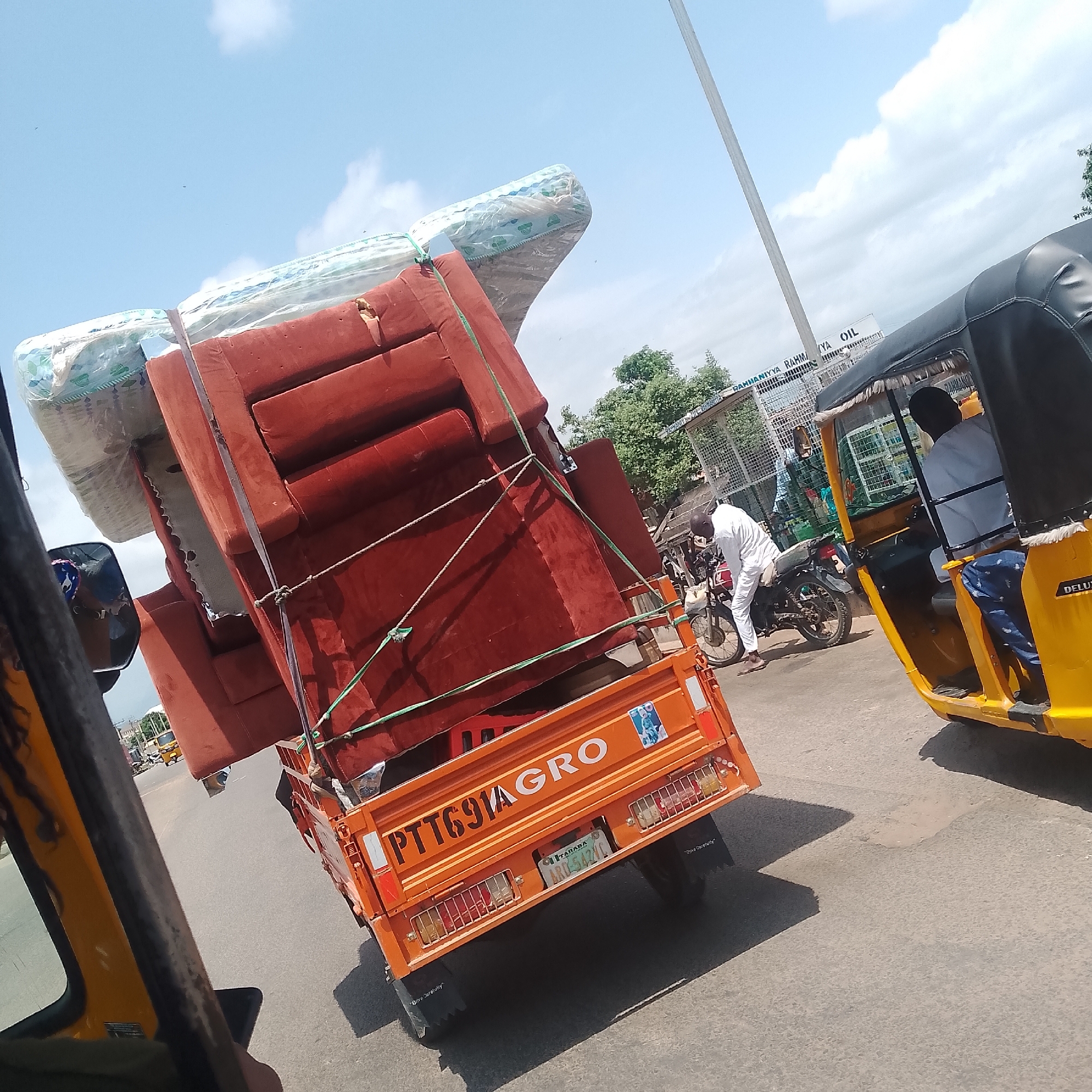 This media file depicts a scene on a street in Jalingo, Nigeria. The primary focus is on an orange motorized tricycle loaded with furniture, including what appears to be a sofa and mattresses wrapped in plastic. The tricycle's license plate is visible, reading "TARABA ARD-5424C," and it also displays "PTT691AGRO" on its rear.
A man in a white garment is seen interacting with a motorcycle parked next to the tricycle. To the right, another similar yellow tricycle is visible, carrying a passenger in the back. The background shows a building with signage that includes "RAHMANIYYA RAHM/YA OIL." Green trees are also present, suggesting an outdoor setting with some vegetation.
The sky is a light blue with scattered clouds, indicating it is daytime. The overall mood appears to be neutral, focusing on the transportation of goods and daily street activity. The tricycle's bumper has the text "Drive Carefully" inscribed on it, a common safety reminder. The image captures a snapshot of local transportation and commerce in Nigeria.