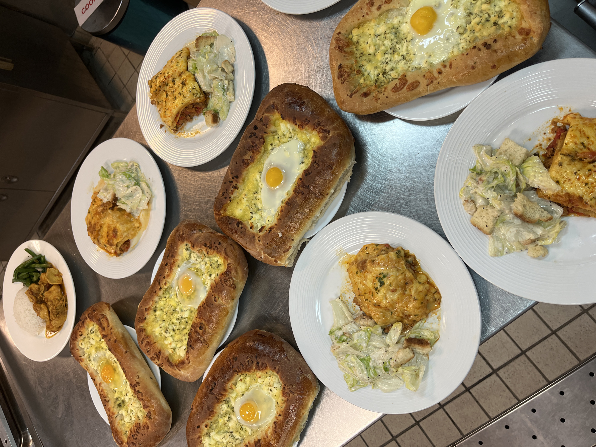 The image displays a top-down view of multiple prepared food plates arranged on a metallic counter, typical of a commercial kitchen, cafeteria, or food service area. The scene highlights a variety of dishes ready for serving.
A central feature of the image is several boat-shaped baked goods, which strongly resemble Khachapuri (Georgian cheese bread). Each piece has a golden-brown, crusty bread exterior, generously filled with melted white cheese and herbs, topped with a runny, bright orange egg yolk in the center. At least five of these unique bread dishes are visible, some fully in view and others partially cropped.
Alongside these baked breads, there are several plates featuring what appears to be a cheesy casserole or baked pasta dish, like lasagna or moussaka, distinguished by its golden, bubbly cheese topping. Each of these servings is accompanied by a side of green salad, which looks like a Caesar salad, complete with croutons and a creamy dressing.
One plate shows a different meal combination: fluffy white rice, a portion of a rich, stew-like dish with pieces of chicken or meat in a savory, orange-hued sauce, and a side of steamed green beans.
All the food items are presented on white ceramic plates with subtle ridged edges. The metallic counter surface reflects the overhead lighting of the indoor setting. In the upper left background, a dark counter or cabinet is visible, along with a dark teal container featuring a white label with red text. The partially visible text reads "COOKI". No people are present in the image, focusing entirely on the array of prepared meals.