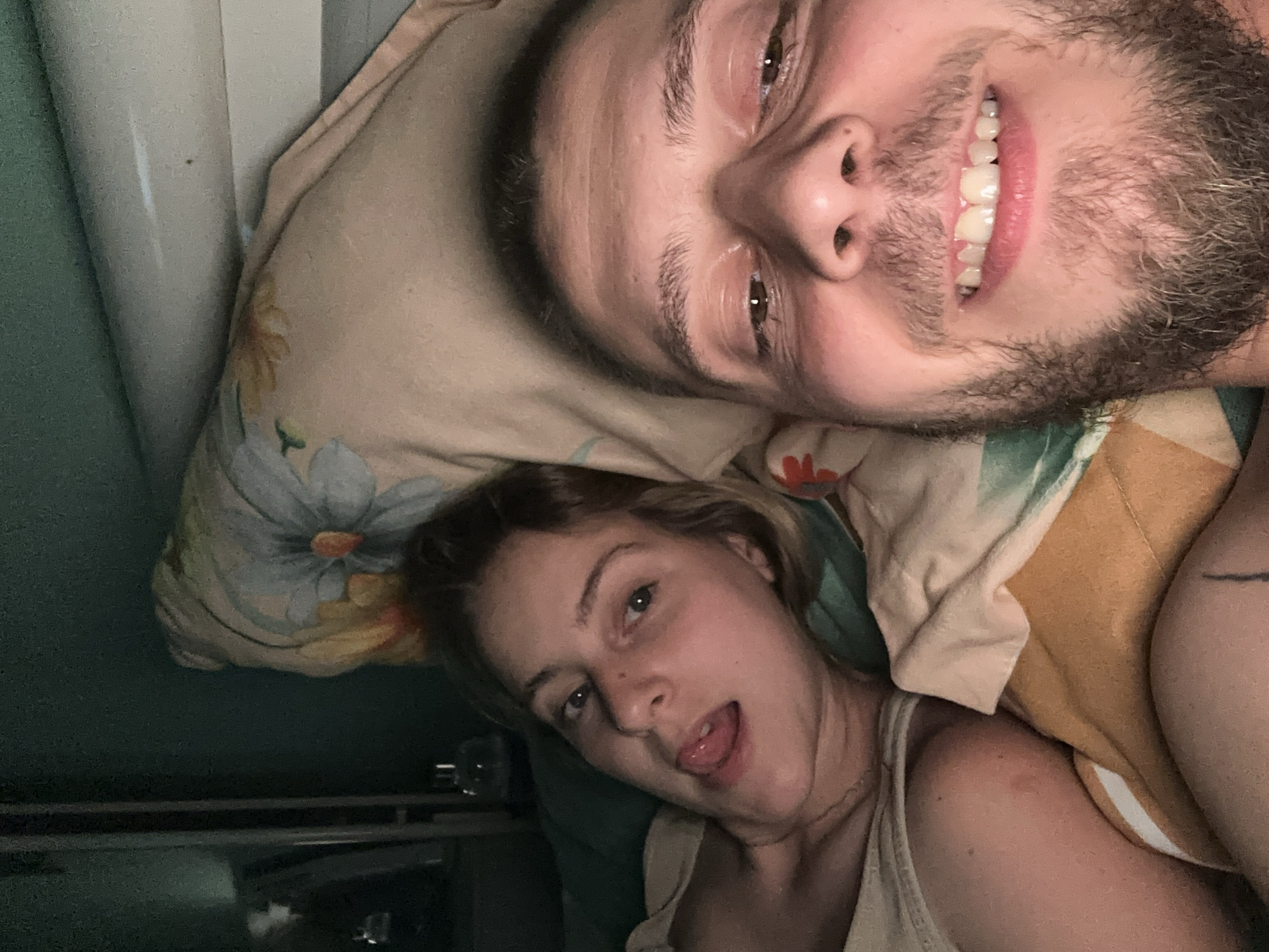 In an indoor scene, a young woman with blonde hair and her boyfriend with a beard are lying in bed. The woman is on the left, wearing a light-colored top, with her tongue sticking out. The man is on the right, with his face close to the camera, smiling. They are both on a bed with floral patterned pillows and bedding. The background is dark green.